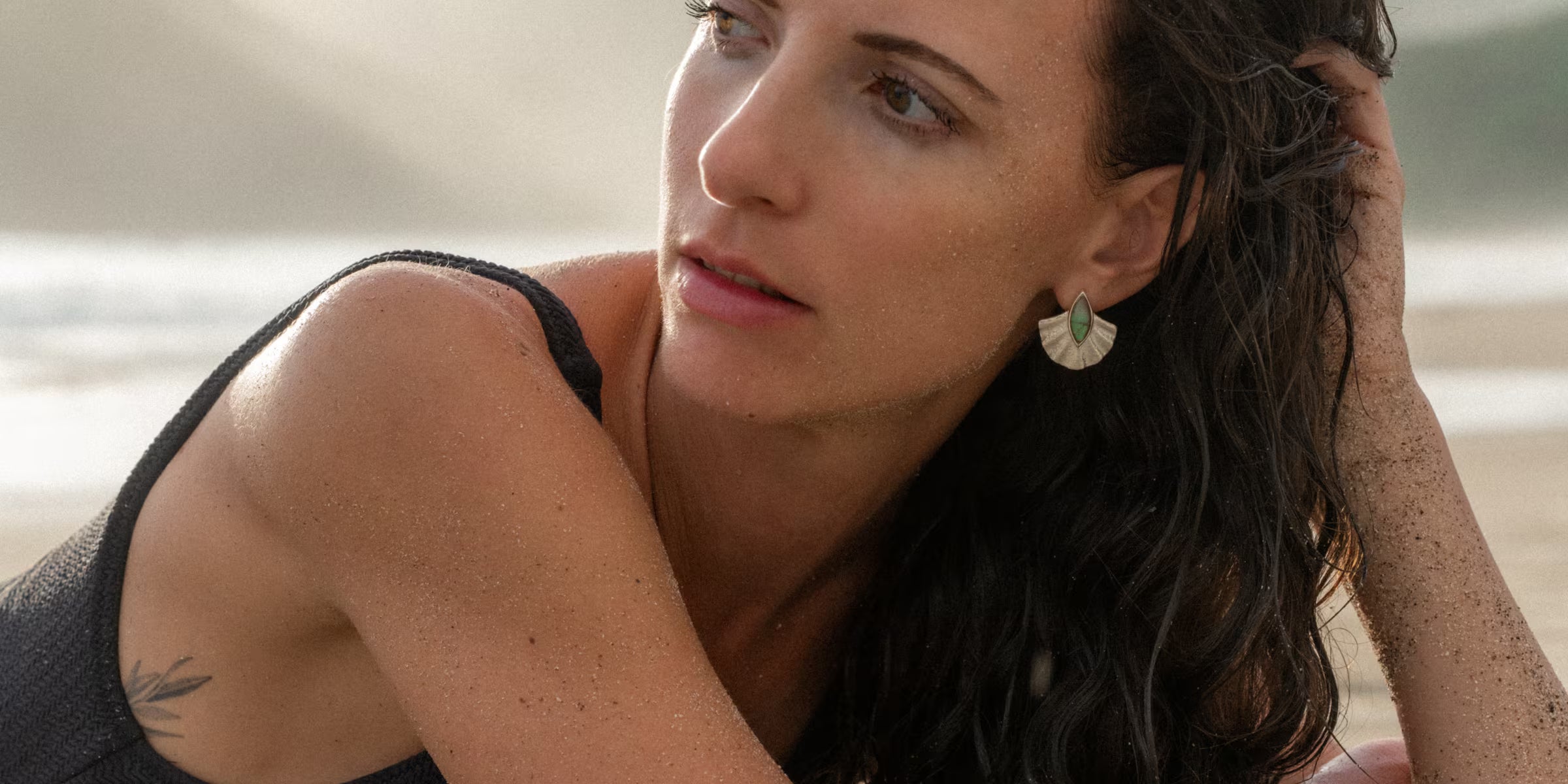 Nahaufnahme eines Models bei einem entspannten Strandtag, das elegante Alun-Ohrringe von der Schmuckmarke ILUH präsentiert. Die fächerartigen Ohrhänger aus recyceltem 925 Sterling Silber sind in Bali handgefertigt und mit einem edlen blaugrünen Labradorit Edelstein besetzt. Sie trägt ein schlichtes schwarzes Oberteil, das die natürliche Eleganz des nachhaltigen Schmucks und den authentischen Beach Vibe des Fotos unterstreicht.