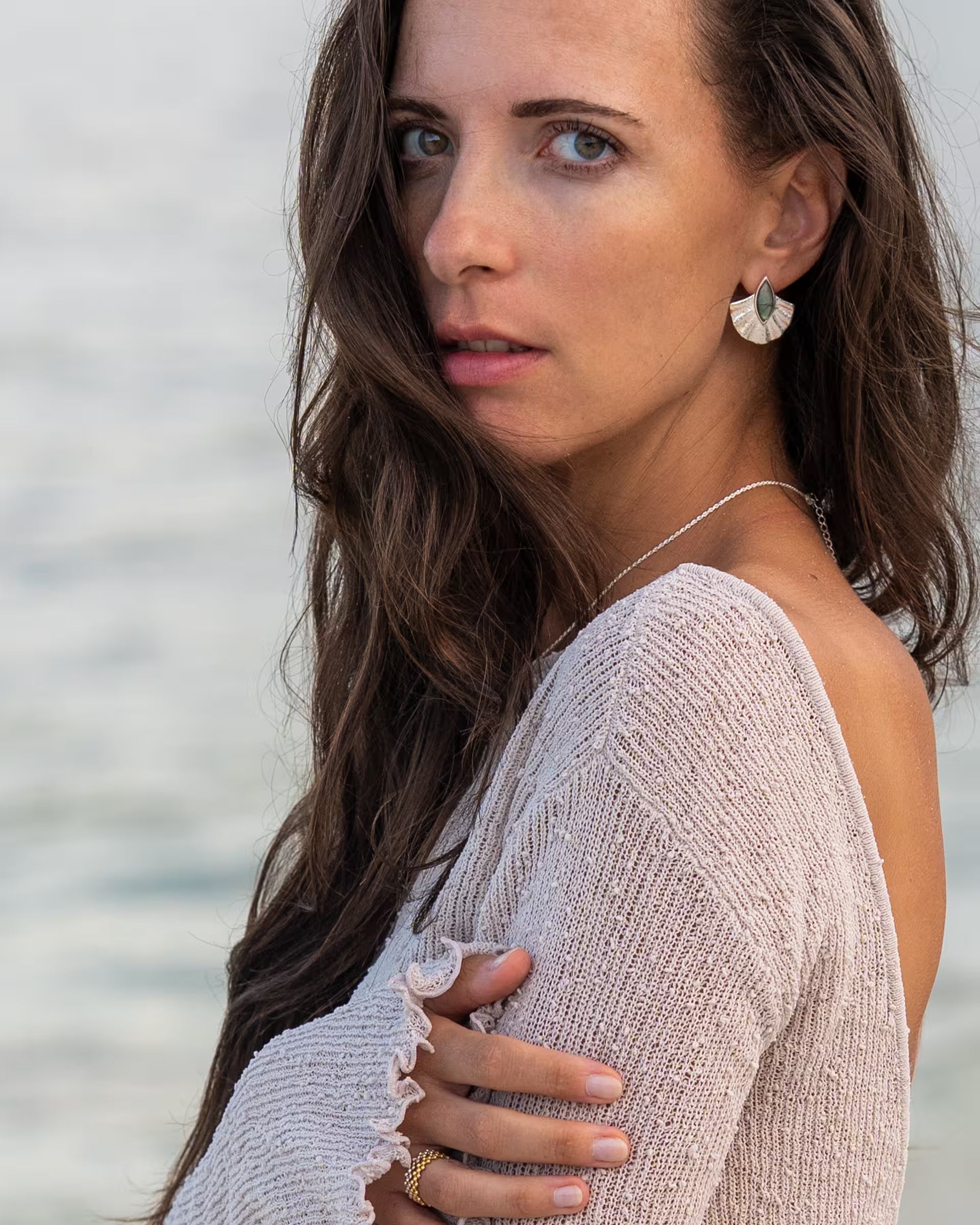 Elegante Nahaufnahme im sommerlichen Look von einer Frau mit langen braunen Haaren im hellen, rückenfreien Strickpullover, die über die Schulter blickt und die auf Bali handgefertigten silbernen ALUN SHELL OHRRINGE mit fächerförmigem Anhänger, verziert mit einem schimmernden, blaugrünen Labradorit-Edelstein, aus 925er Silber von der Schmuckmarke ILUH trägt, vor dem Hintergrund des Meeres.