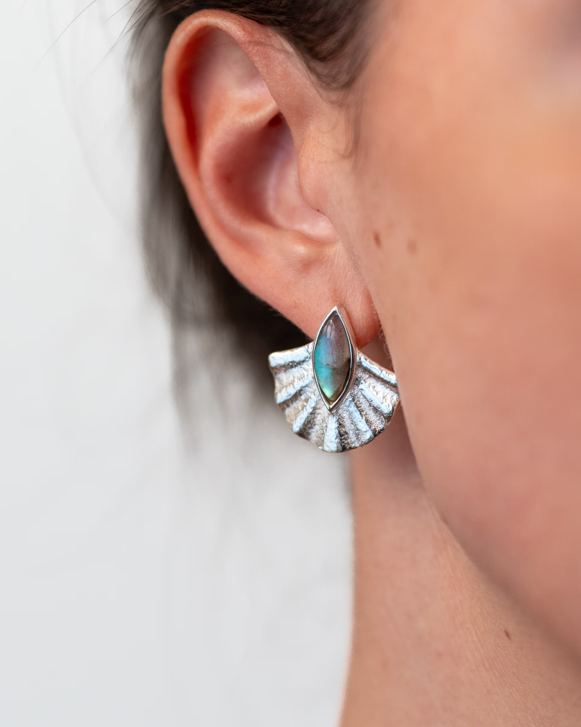 ALUN SHELL LABRADORITE OHRRINGE SILBER, silberner Schmuck von der Schmuckmarke ILUH, handgefertigt auf Bali – Stilvolle Nahaufnahme im Profil von dem Ohr einer Frau, die fächerförmige Ohrringe aus 925er Silber mit einem intensiv blaugrün schimmernden Labradorit-Edelstein im Cabochonschliff vor einem hellen, neutralen Hintergrund präsentiert.