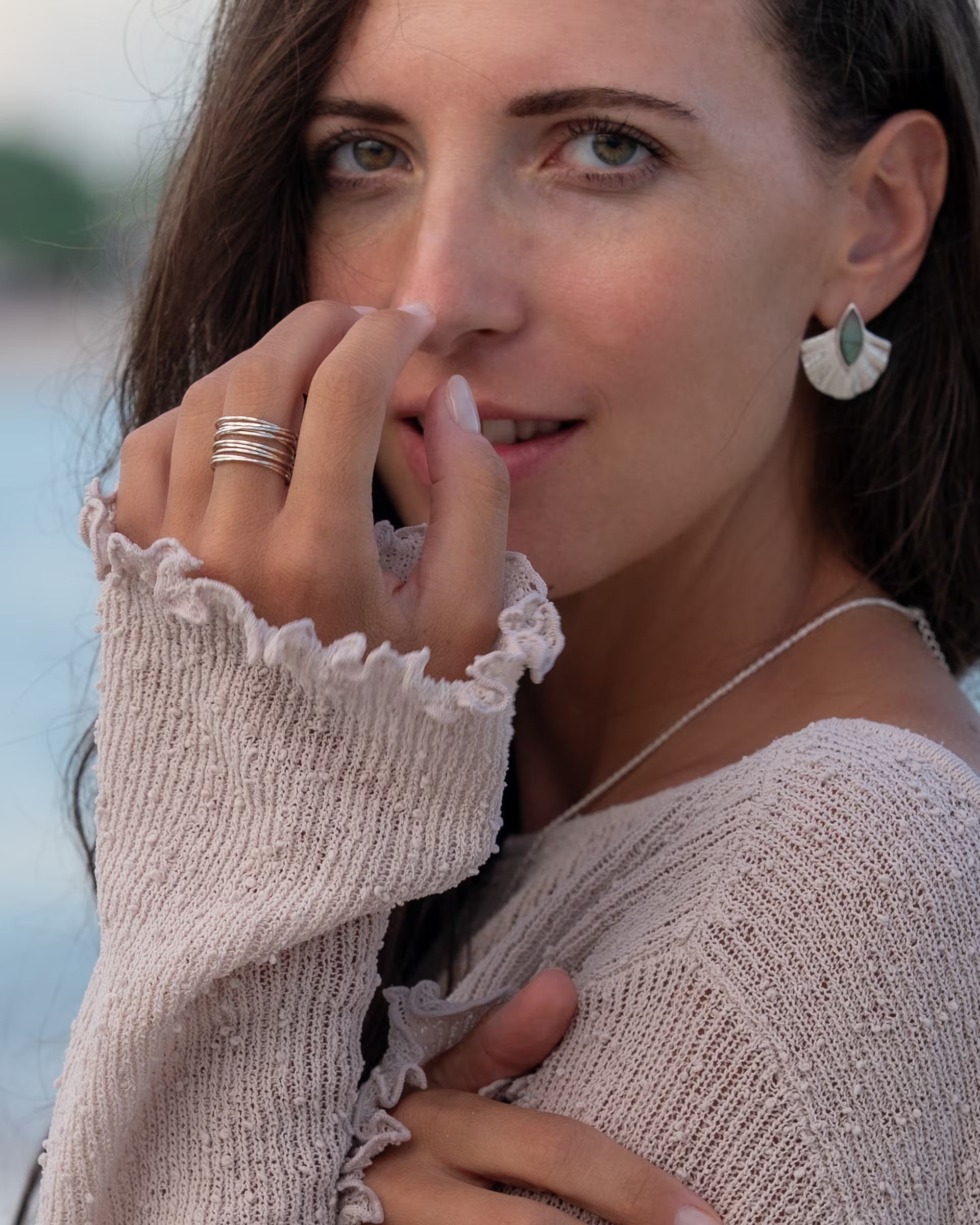 Elegante Nahaufnahme im herbstlich-sommerlichen Look von einer Frau mit langen braunen Haaren im hellen Grobstrickpullover, die eine Hand zart an das Gesicht legt und die auf Bali handgefertigten silbernen ALUN SHELL OHRRINGE mit fächerförmigem Anhänger und blaugrünem Labradorit-Edelstein sowie den mehrlagigen AURA RING im Stacking-Look aus 925er Silber von der Schmuckmarke ILUH zeigt.