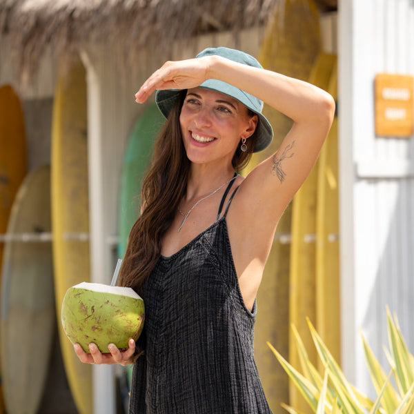 Timea, Gründerin des Schmuck-Labels ILUH, mit entspanntem Surfer-Vibe vor einer Kulisse aus Surfbrettern und mit einem frischen Kokosnuss in der Hand; das Bild unterstreicht den natürlichen, handgefertigten Charakter des minimalistischen Schmucks aus Bali.