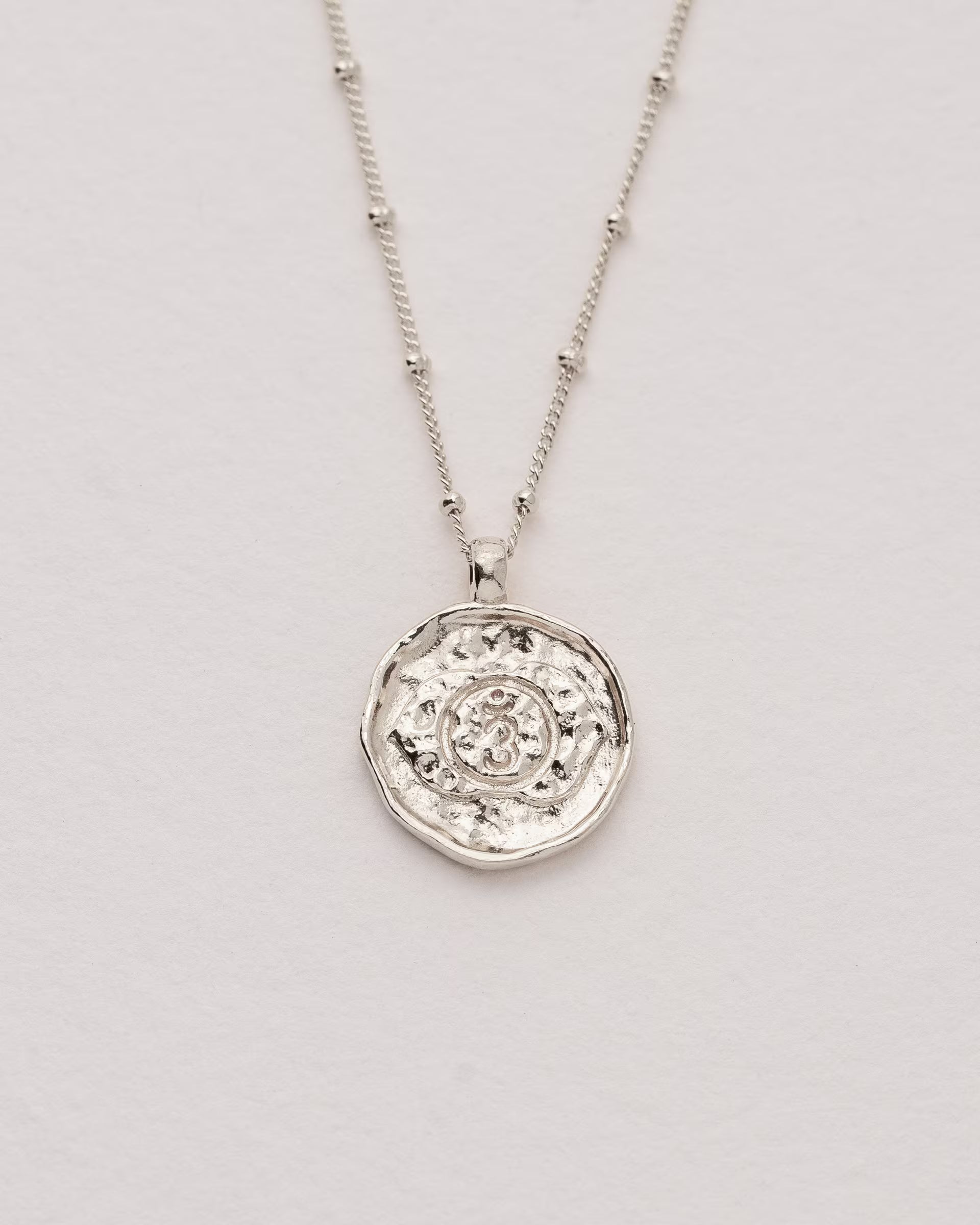 #Motiv_Ajna CHAKRA AJNA NECKLACE SILBER, silberne Halskette von ILUH, handgefertigt auf Bali – Spirituelles Produktfoto im minimalistischen Boho-Look von einer feinen Satellitenkette aus 925er Silber mit einem runden Münzanhänger, der das detailliert geprägte Ajna-Stirnchakra-Symbol für Intuition zeigt, flach präsentiert auf einem hellen, neutralen Untergrund.