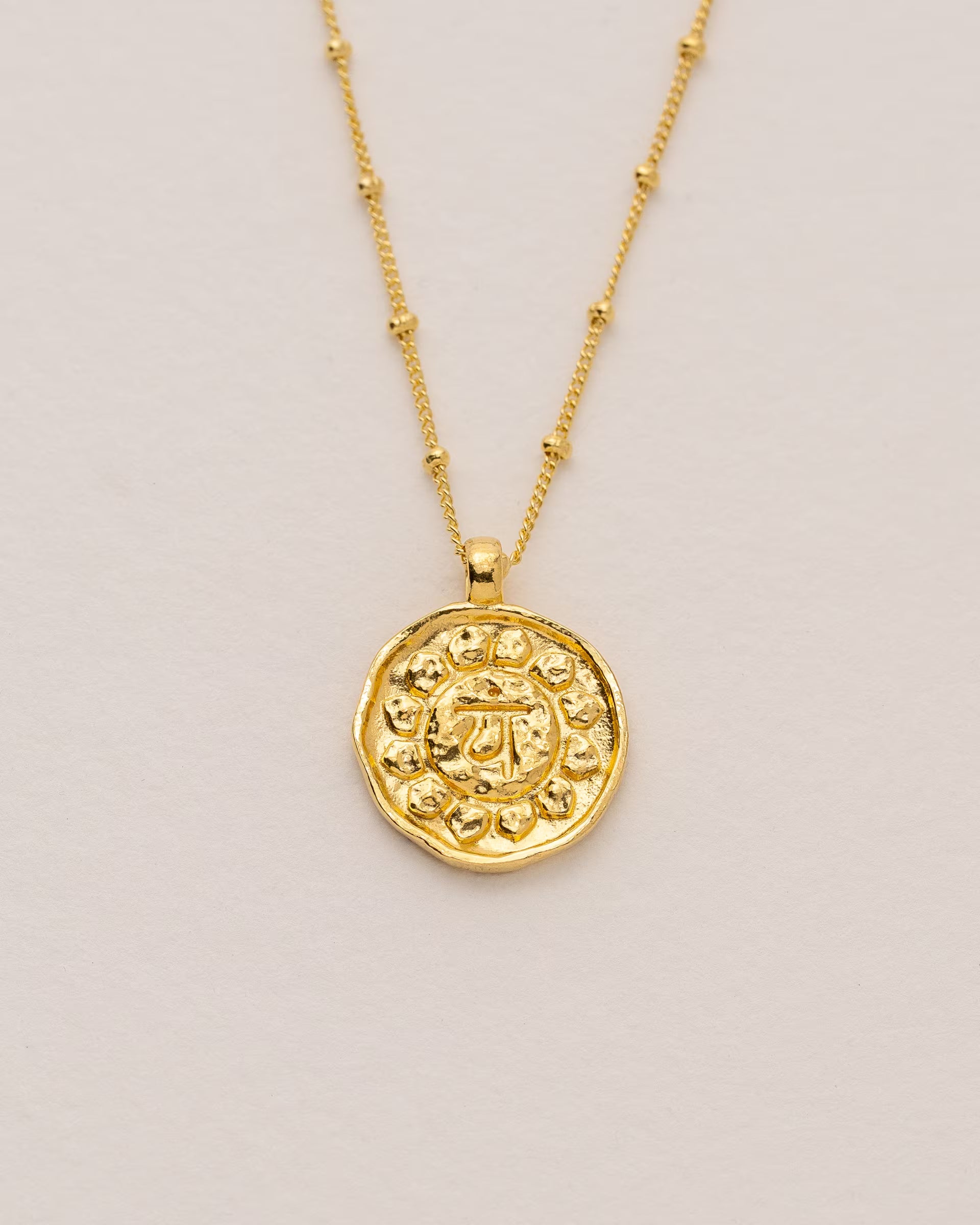 #Motiv_Anahata CHAKRA ANAHATA NECKLACE GOLD, goldene Halskette von ILUH, handgefertigt auf Bali – Spirituelles Produktfoto im minimalistischen Boho-Look von einer feinen Satellitenkette aus 18k vergoldetem 925er Silber mit einem runden Münzanhänger, der das detailliert geprägte Anahata-Herzchakra-Symbol zeigt, flach präsentiert auf einem hellen, neutralen Untergrund.