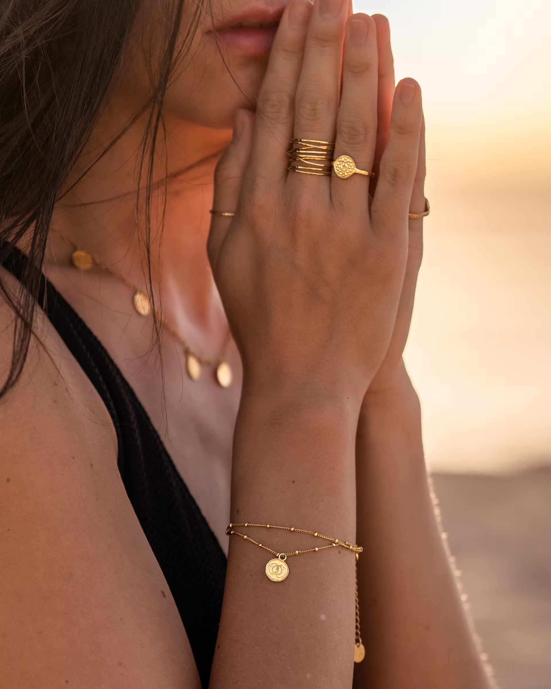 CHAKRA ARMBAND UND RINGE, goldener Schmuck von ILUH, handgefertigt auf Bali – Sinnliche Nahaufnahme am sommerlichen Strand von den zum Gebet gefalteten Händen einer Frau, die das filigrane, zweireihige Chakra-Armband am Handgelenk, den runden CHAKRA RING sowie den breiten, mehrreihigen, gehämmerten AURA RING im Wickeldesign aus 18k vergoldetem 925er Silber im goldenen Licht des Sonnenuntergangs präsentiert.