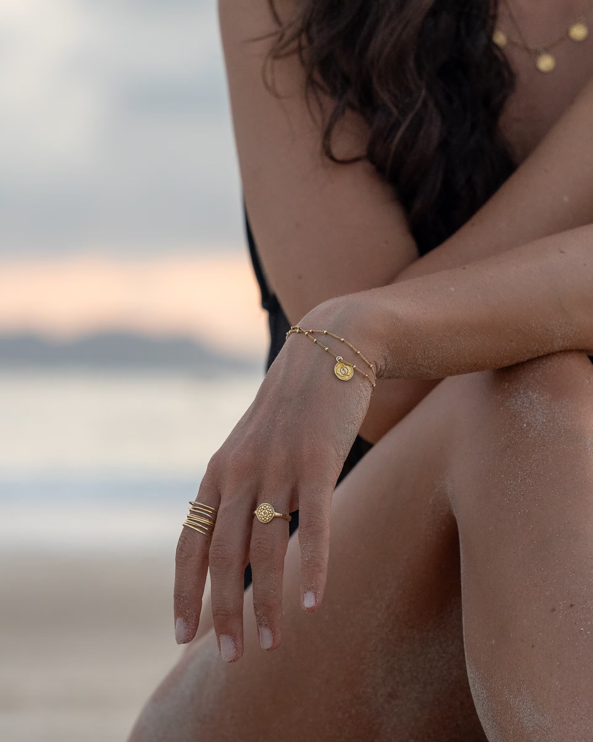 CHAKRA SCHMUCKSET, goldene Halskette, Ohrringe und Armband von ILUH, handgefertigt auf Bali – Stimmungsvolle Nahaufnahme im spirituellen Beach-Look von einer Frau im schwarzen Badeanzug am Strand, die im sanften Licht des Sonnenuntergangs die PRANA KETTE mit sieben Chakra-Münzanhängern, die passenden CHAKRA OHRRINGE als Creolen sowie das feine Armband und Ringe aus 18k vergoldetem 925er Silber präsentiert.