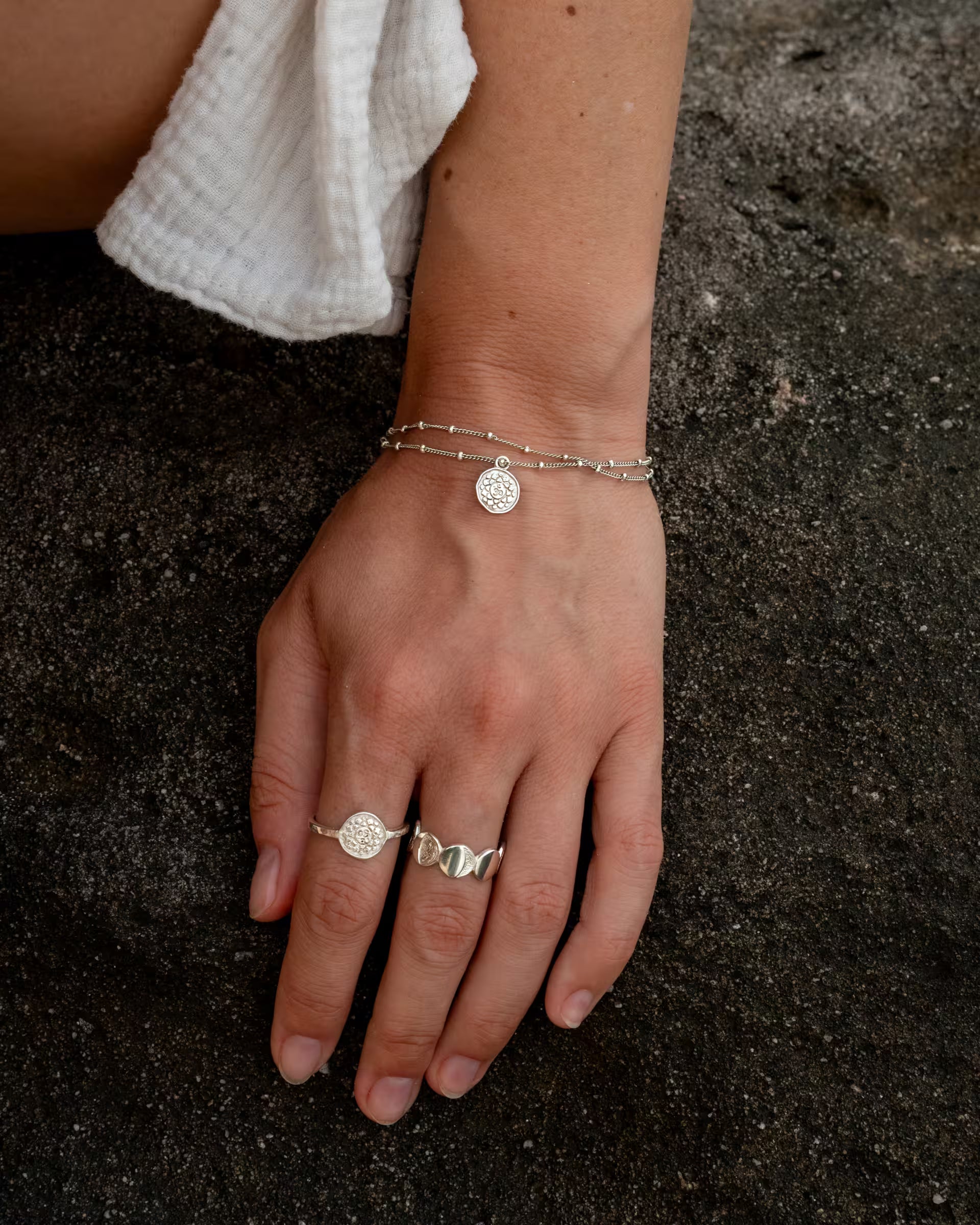 Nahaufnahme im spirituellen Beach-Look von einer Hand auf dunklem Vulkansand, die eine Kombination aus auf Bali handgefertigtem silbernem Schmuck aus der Chakra-Kollektion – bestehend aus dem dopplagigen CHAKRA ARMBAND mit feiner Satellitenkette und rundem Motiv-Anhänger sowie dem passenden CHAKRA RING mit geprägtem Motiv am Zeigefinger, sowie den offenen CHANDRA RING – aus 925er Silber von der Schmuckmarke ILUH trägt.