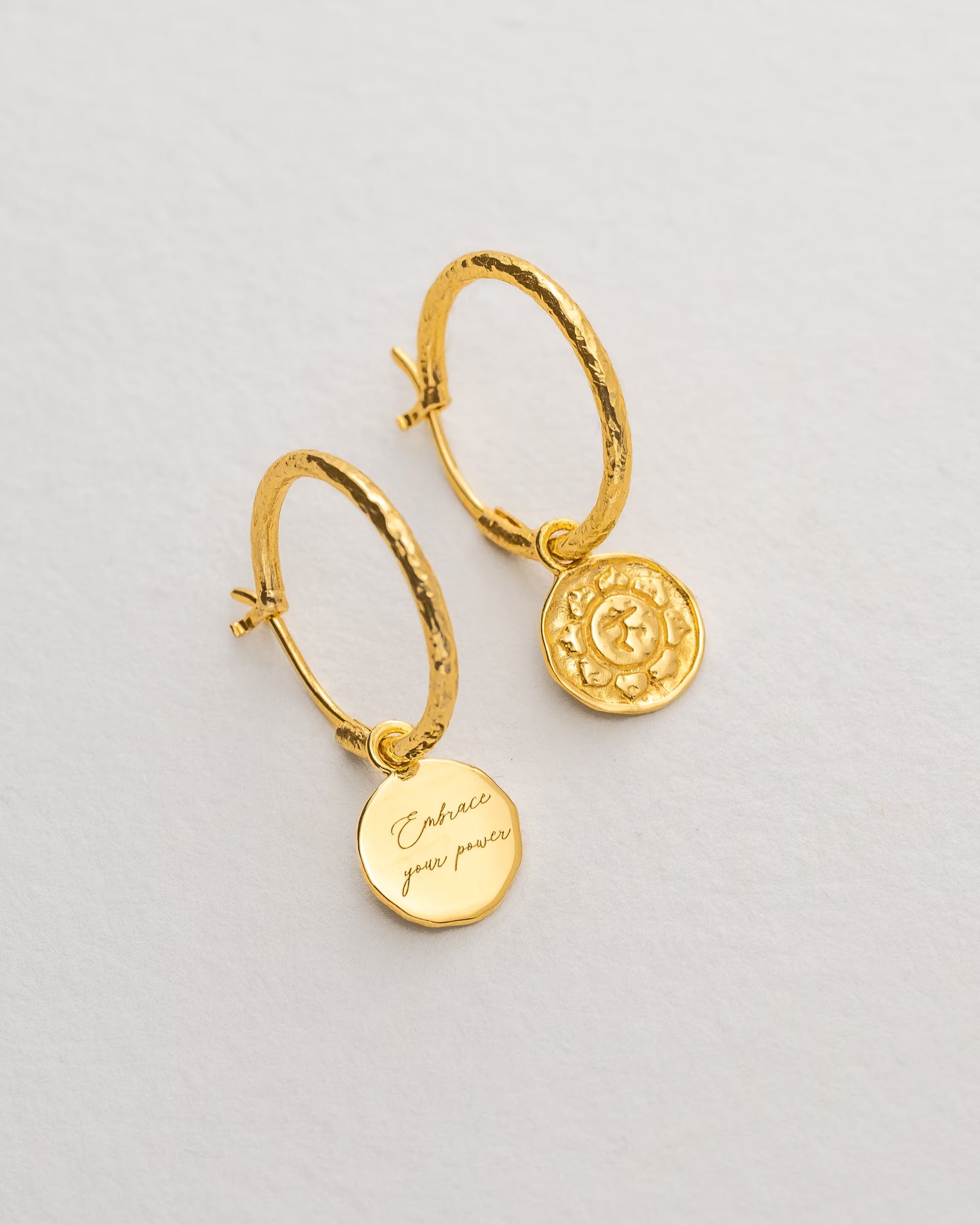 Filigrane ILUH Creolen aus nachhaltigem, recyceltem Silber mit einer hochwertigen 18k Goldplattierung und einem runden Anhänger mit Solarplexus-Chakra-Motiv. Das Foto ist eine präzise Nahaufnahme auf strukturiertem Büttenpapier, die durch das weiche Licht die organische Textur des Schmucks sowie die zarte Gravur mit der Intention 'Embrace your power' hervorhebt – ein zeitloses Beispiel für hochwertigen, spirituellen Schmuck mit tiefer Bedeutung.