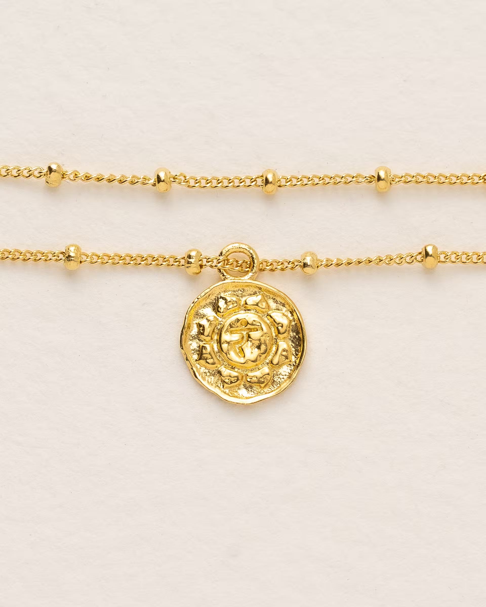 #Motiv_Manipura CHAKRA MANIPURA ARMBAND GOLD, goldenes Armband von ILUH, handgefertigt auf Bali – Spirituelles Produktfoto im minimalistischen Boho-Look von einem feinen Satellitenarmband aus 18k vergoldetem 925er Silber mit einem runden Münzanhänger, der das originale Sanskrit-Zeichen für das Manipura-Solarplexuschakra, eingebettet in kunstvolle Lotusblütenblätter zur Stärkung des Energiezentrums, zeigt, flach präsentiert auf einem hellen, neutralen Büttenpapier-Untergrund.