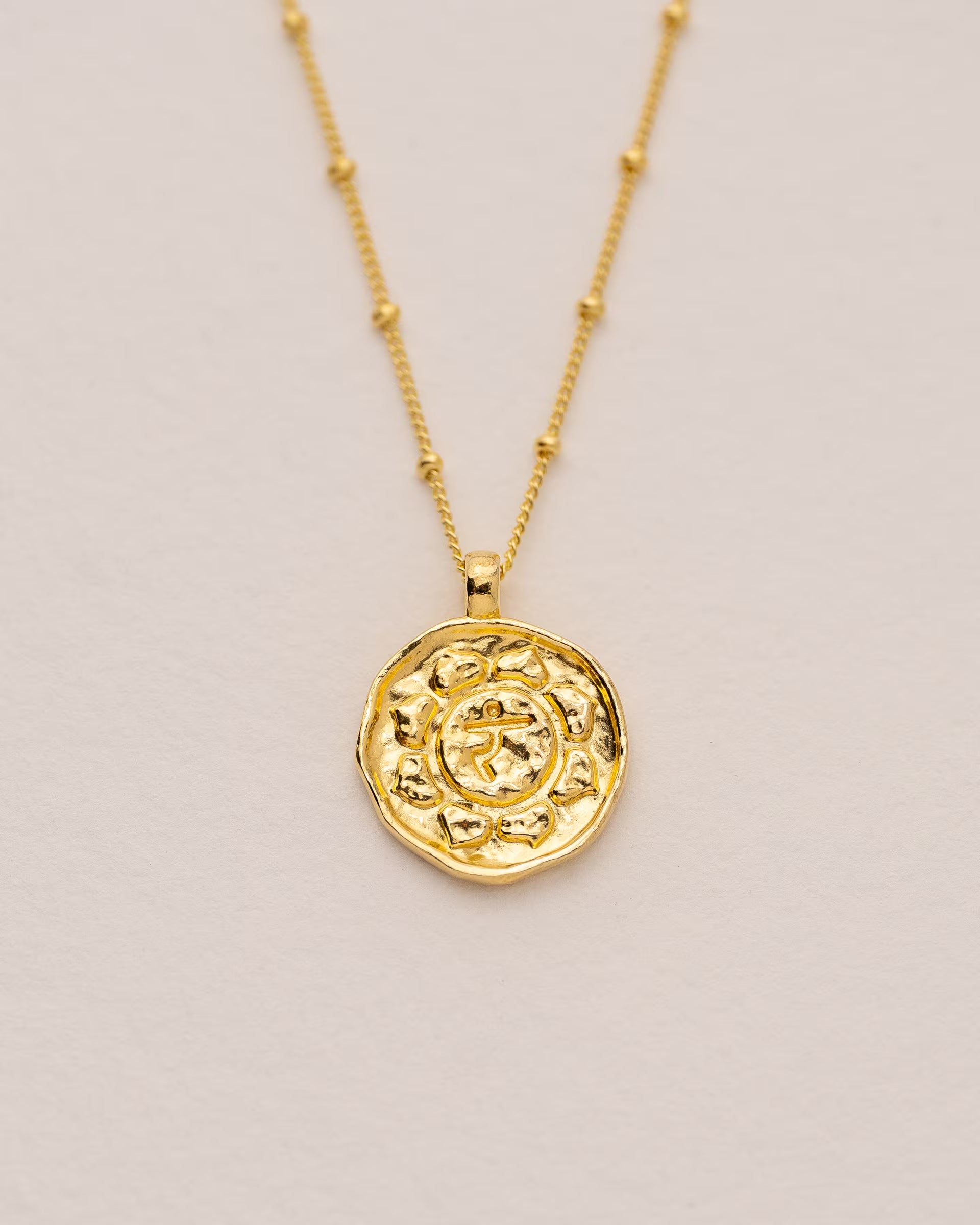 #Motiv_Manipura CHAKRA MANIPURA NECKLACE GOLD, goldene Halskette von ILUH, handgefertigt auf Bali – Spirituelles Produktfoto im minimalistischen Boho-Look von einer feinen Satellitenkette aus 18k vergoldetem 925er Silber mit einem runden Münzanhänger, der das originale Sanskrit-Zeichen für das Manipura-Solarplexuschakra, eingebettet in kunstvolle Lotusblütenblätter zur Stärkung des Energiezentrums, zeigt, flach präsentiert auf einem hellen, neutralen Büttenpapier-Untergrund.