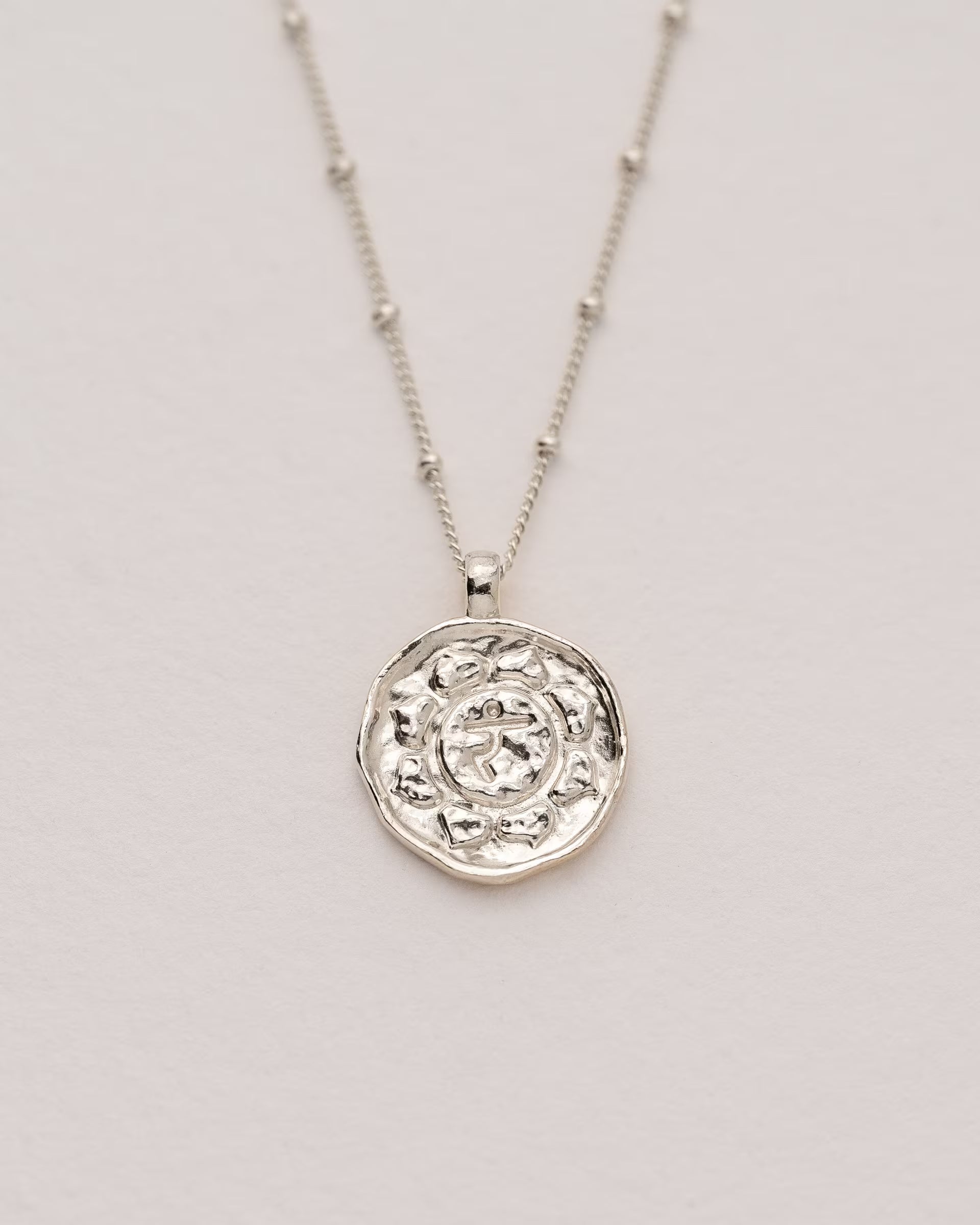 #Motiv_Manipura CHAKRA MANIPURA NECKLACE SILBER, silberne Halskette von ILUH, handgefertigt auf Bali – Spirituelles Produktfoto im minimalistischen Boho-Look von einer feinen Satellitenkette aus 925er Silber mit einem runden Münzanhänger, der das originale Sanskrit-Zeichen für das Manipura-Solarplexuschakra, eingebettet in kunstvolle Lotusblütenblätter zur Stärkung des Energiezentrums, zeigt.