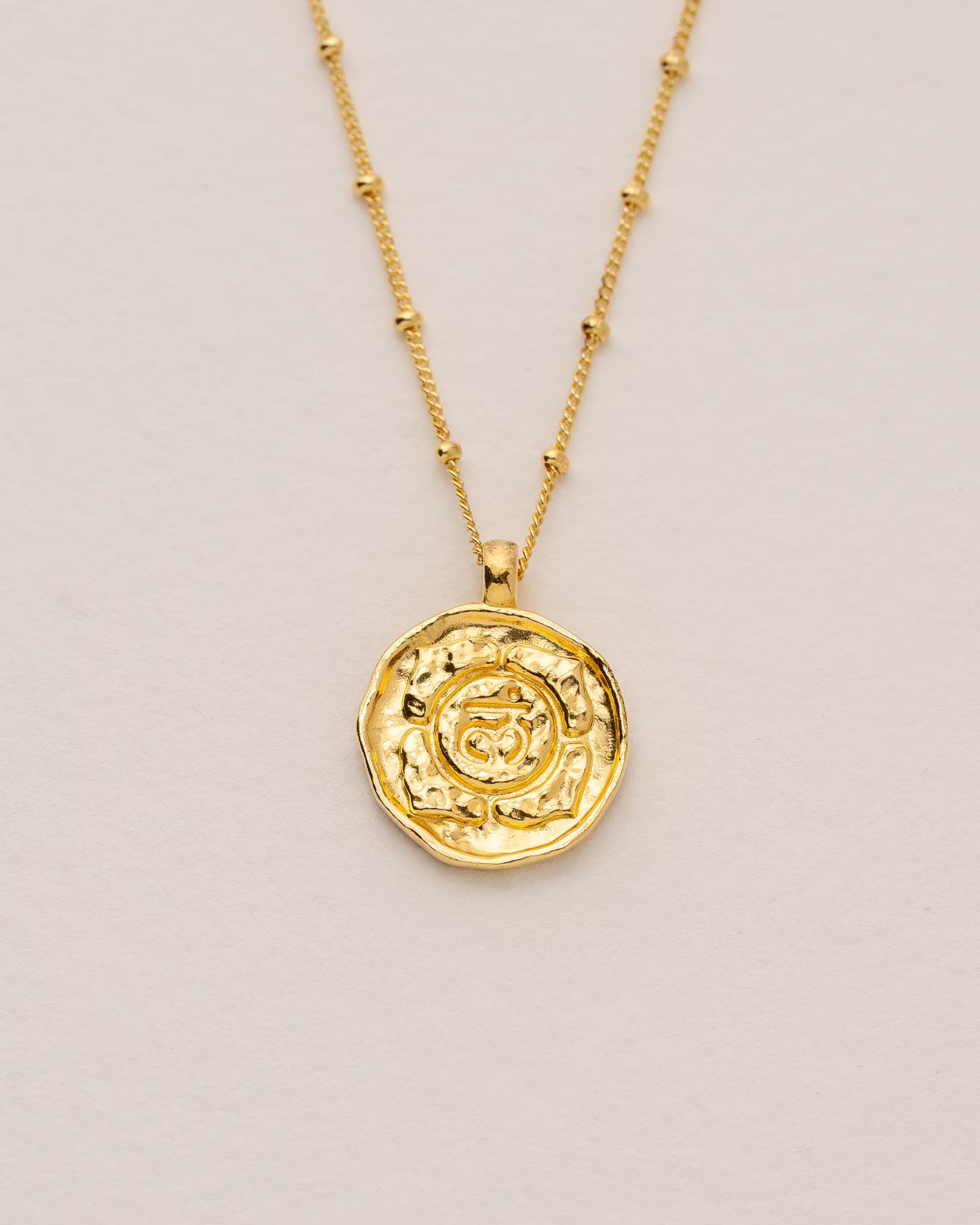 #Motiv_Muladhara CHAKRA MULADHARA NECKLACE GOLD, goldene Halskette von ILUH, handgefertigt auf Bali – Spirituelles Produktfoto im minimalistischen Boho-Look von einer feinen Satellitenkette aus 18k vergoldetem 925er Silber mit einem runden Münzanhänger, der das originale Sanskrit-Zeichen für das Wurzelchakra eingebettet in kunstvolle Lotusblütenblätter zeigt, um die Stärkung des Energiezentrums zu fördern, flach präsentiert auf einem hellen, neutralen Büttenpapier-Untergrund.