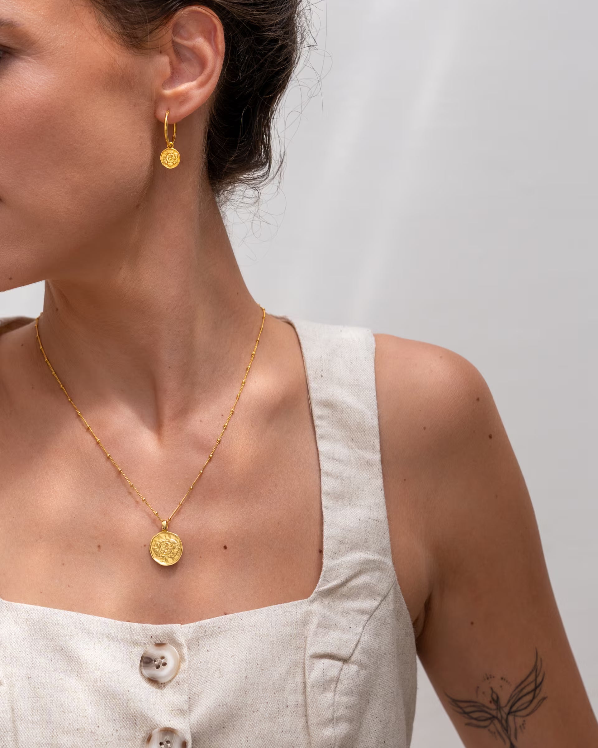 CHAKRA SCHMUCKSET, goldene Halskette und Ohrringe von ILUH, handgefertigt auf Bali – Elegante Nahaufnahme von einer Frau im hellen Leinen-Oberteil im Profil vor einer hellen Wand, die eine feine Satellitenkette mit rundem, geprägtem Chakra-Münzanhänger sowie die passenden CHAKRA OHRRINGE als kleine Creolen aus 18k vergoldetem 925er Silber präsentiert.
