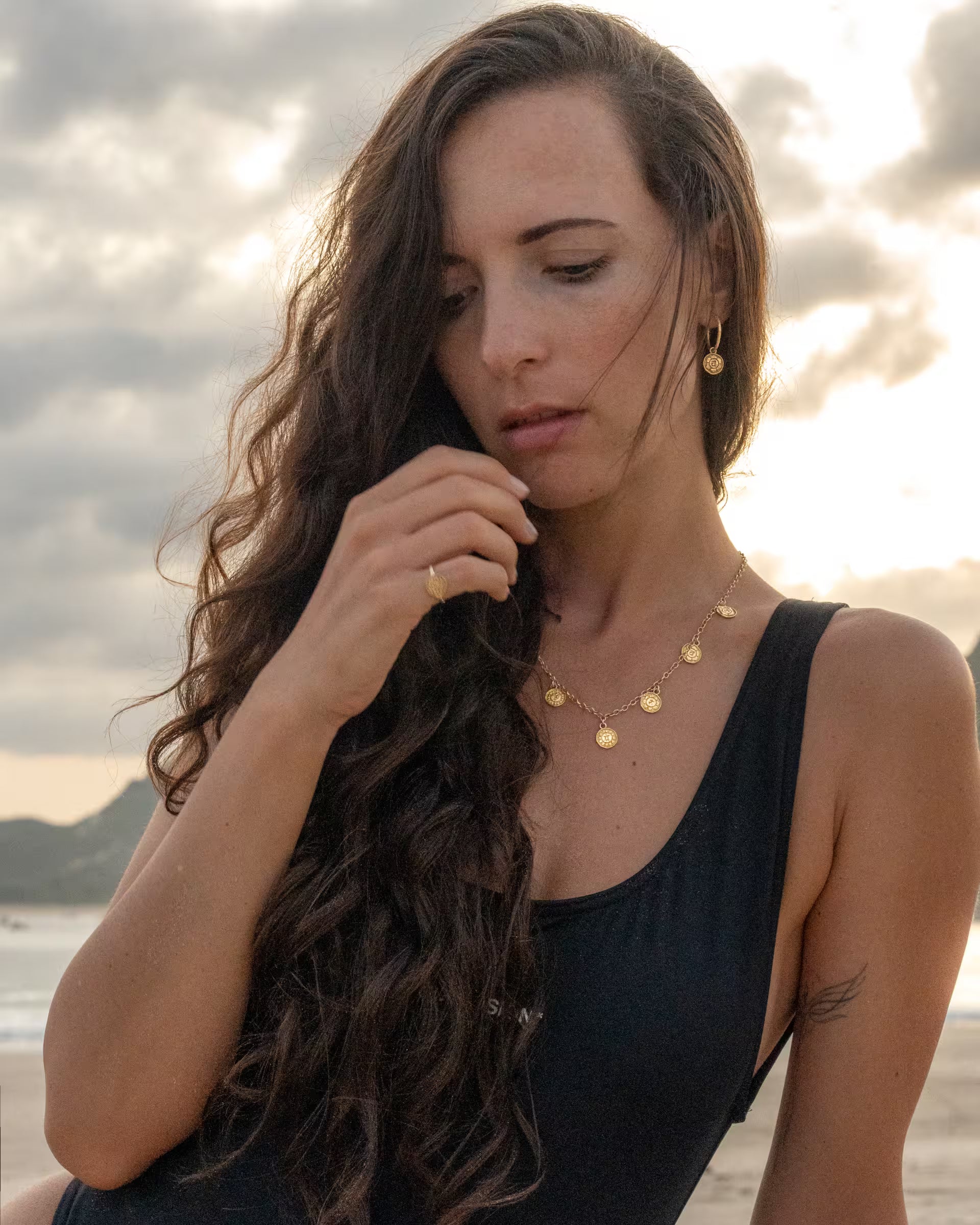 Stimmungsvolle Nahaufnahme im spirituellen Beach-Look von einer Frau mit langen, lockigen Haaren im schwarzen Badeanzug am Strand, die den Blick senkt und eine Kombination aus auf Bali handgefertigtem goldenem Schmuck aus der Chakra-Kollektion – bestehend aus der PRANA KETTE mit sieben runden Chakra-Motiven als Anhänger, den passenden CHAKRA OHRRINGEN sowie einem feinen CHAKRA RING – aus 18k vergoldetem 925er Silber von der Schmuckmarke ILUH trägt.
