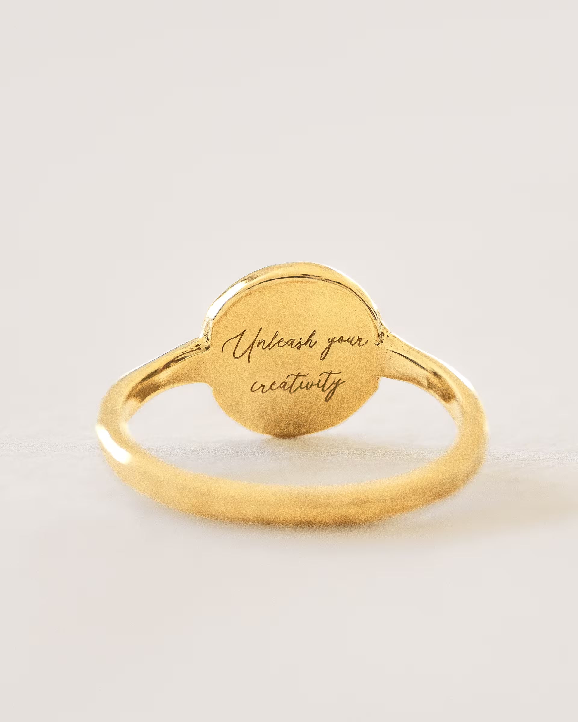 Filigraner ILUH Ring aus 18k vergoldetem, recyceltem Silber mit einer runden Platte; Nahaufnahme der Innenseite mit der eingravierten Intention 'Unleash your creativity', präsentiert auf elegantem, strukturiertem Büttenpapier – ein hochwertiges Schmuckstück, das dem Sakral-Chakra zugeordnet ist und Inspiration sowie künstlerische Entfaltung symbolisiert.