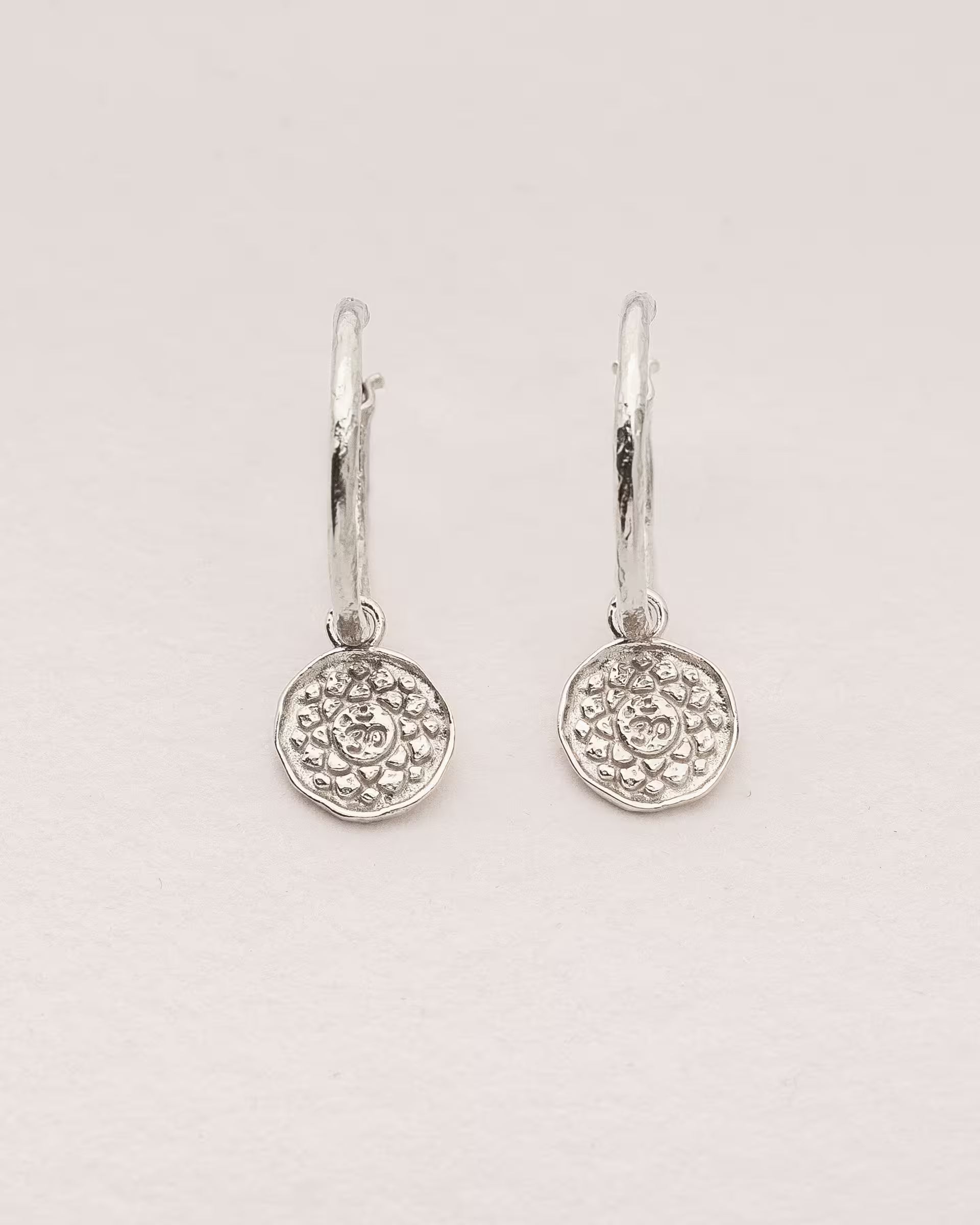 #Motiv_Sahasrara CHAKRA SAHASRARA OHRRINGE SILBER Produktbild – Diese handgefertigten Ohrringe von ILUH aus Bali im minimalistischen Boho-Stil bestehen aus strukturierten Creolen mit einem runden Anhänger, der das originale Sanskrit-Zeichen für das Sahasrara-Kronenchakra zeigt; eingebettet in kunstvolle Lotusblütenblätter dienen sie als kraftvolles Symbol zur Stärkung deines Energiezentrums.