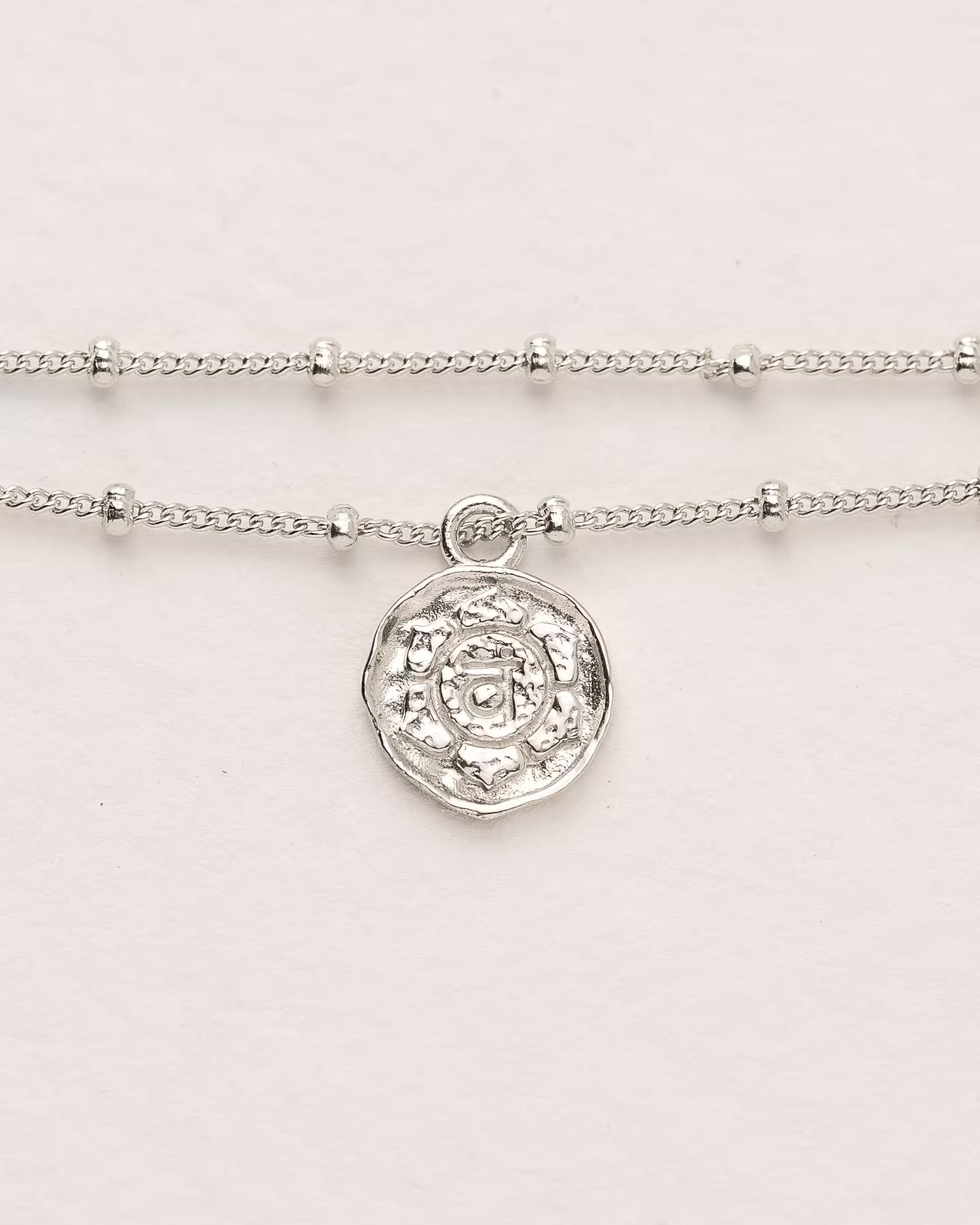 #Motiv_Svadhisthana CHAKRA SVADHISTHANA ARMBAND SILBER, silbernes Armband von ILUH, handgefertigt auf Bali – Spirituelles Produktfoto im minimalistischen Boho-Look von einem feinen, doppelten Satellitenarmband aus 925er Silber mit einem runden Münzanhänger, der das originale Sanskrit-Zeichen für das Svadhisthana-Sakralchakra, eingebettet in kunstvolle Lotusblütenblätter zur Stärkung des Energiezentrums, zeigt.