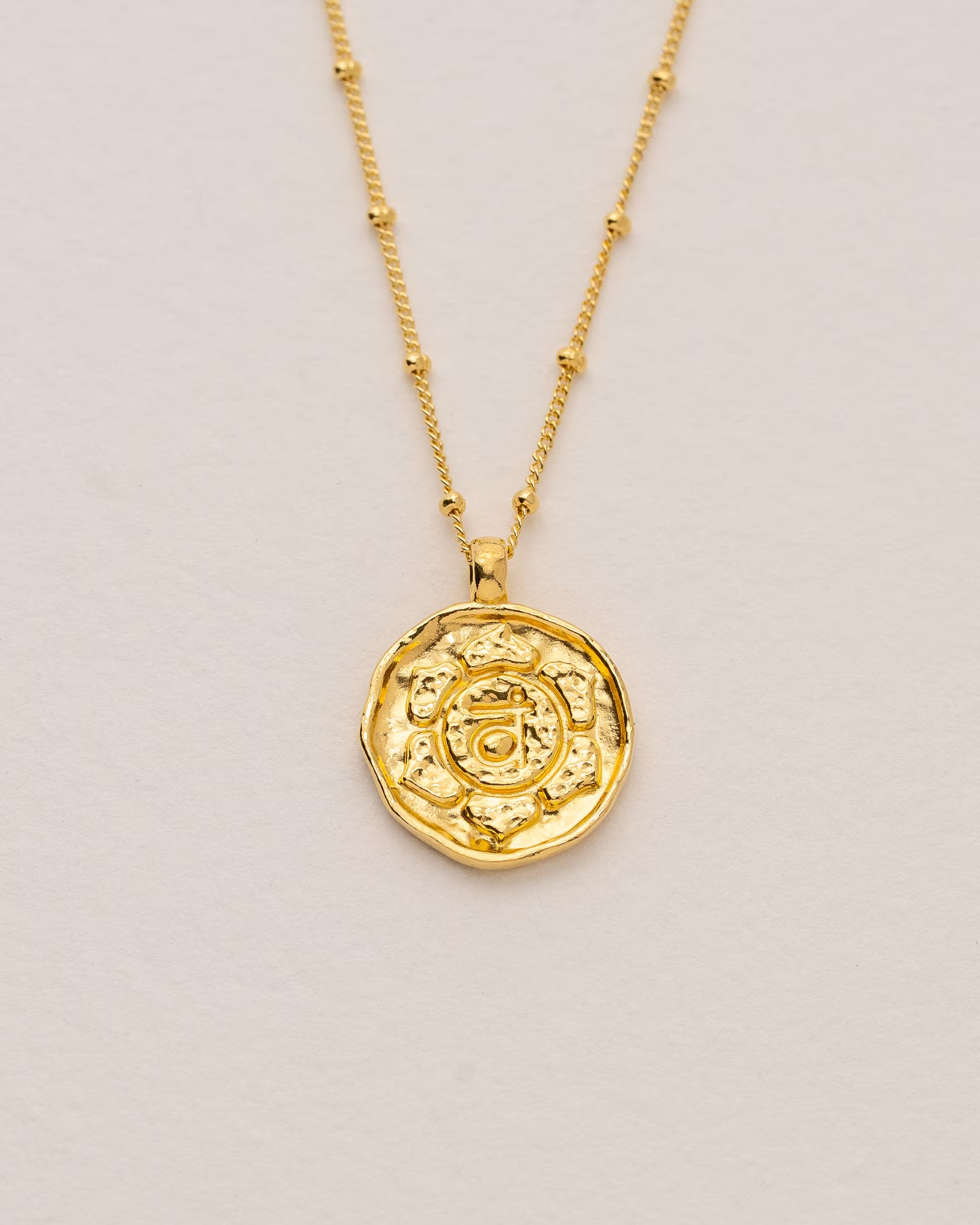 #Motiv_Svadhisthana CHAKRA SVADHISTHANA NECKLACE GOLD, goldene Halskette von ILUH, handgefertigt auf Bali – Spirituelles Produktfoto im minimalistischen Boho-Look von einer feinen Satellitenkette aus 18k vergoldetem 925er Silber mit einem runden Münzanhänger, der das detailliert geprägte Svadhisthana-Sakralchakra als energetisches Energiezentrum zeigt, flach präsentiert auf einem hellen, neutralen Büttenpapier Untergrund.
