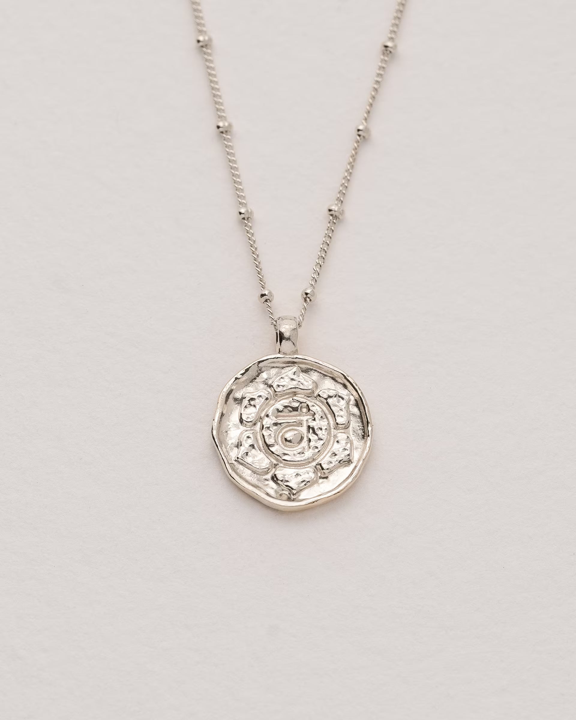 #Motiv_Svadhisthana CHAKRA SVADHISTHANA NECKLACE SILBER, silberne Halskette von ILUH, handgefertigt auf Bali – Spirituelles Produktfoto im minimalistischen Boho-Look von einer feinen Satellitenkette aus 925er Silber mit einem runden Münzanhänger, der das originale Sanskrit-Zeichen für das Svadhisthana-Sakralchakra, eingebettet in kunstvolle Lotusblütenblätter zur Stärkung des Energiezentrums, zeigt.
