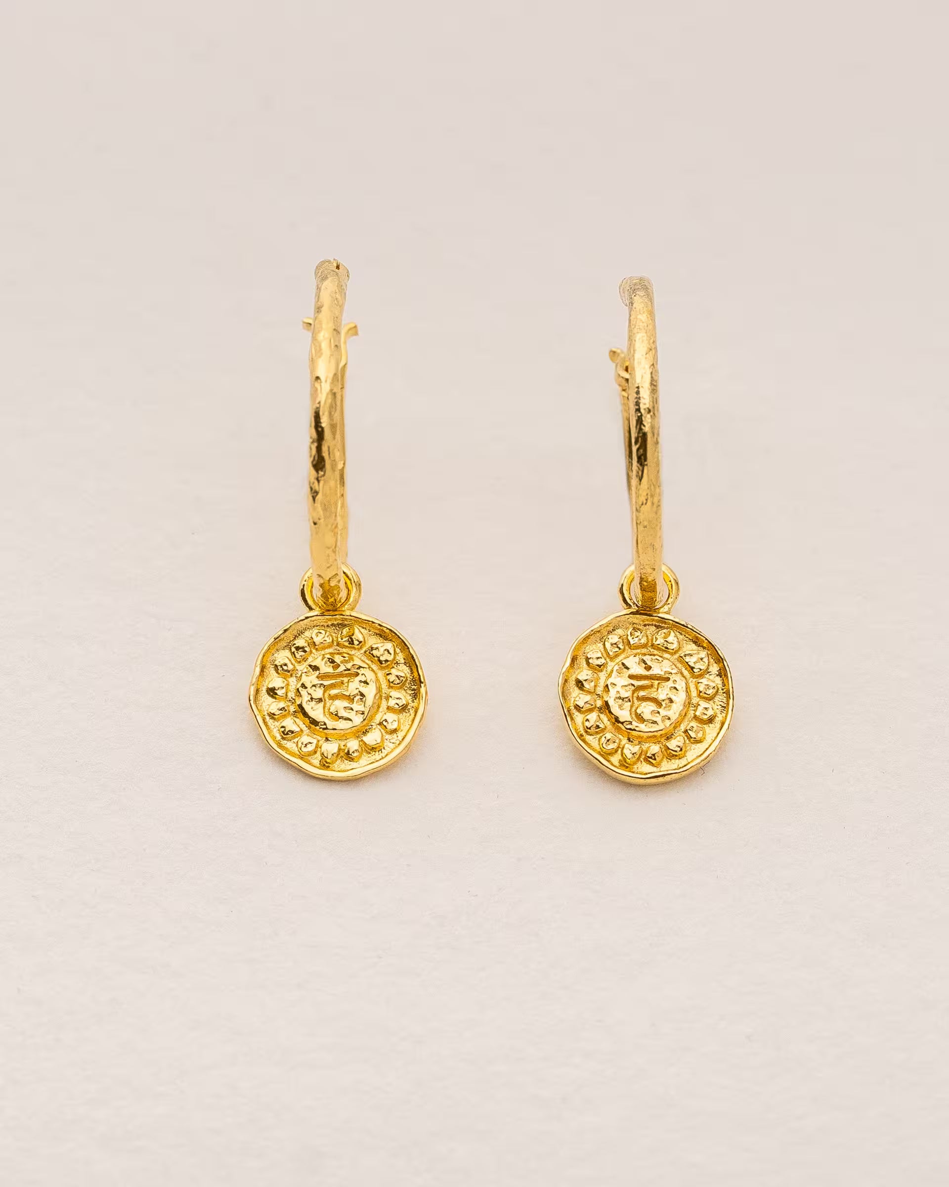 #Motiv_Vishuddha CHAKRA VISHUDDHA OHRRINGE GOLD, kleine goldene Creolen-Ohrringe von ILUH, handgefertigt auf Bali – Elegantes Produktfoto im spirituellen Boho-Look von einem Paar filigraner Creolen aus 18k vergoldetem 925er Silber mit je einem kleinen, runden Anhänger, der das originale Sanskrit-Zeichen für das Vishuddha-Halschakra, eingebettet in kunstvolle Lotusblütenblätter zur Stärkung des Energiezentrums, zeigt.