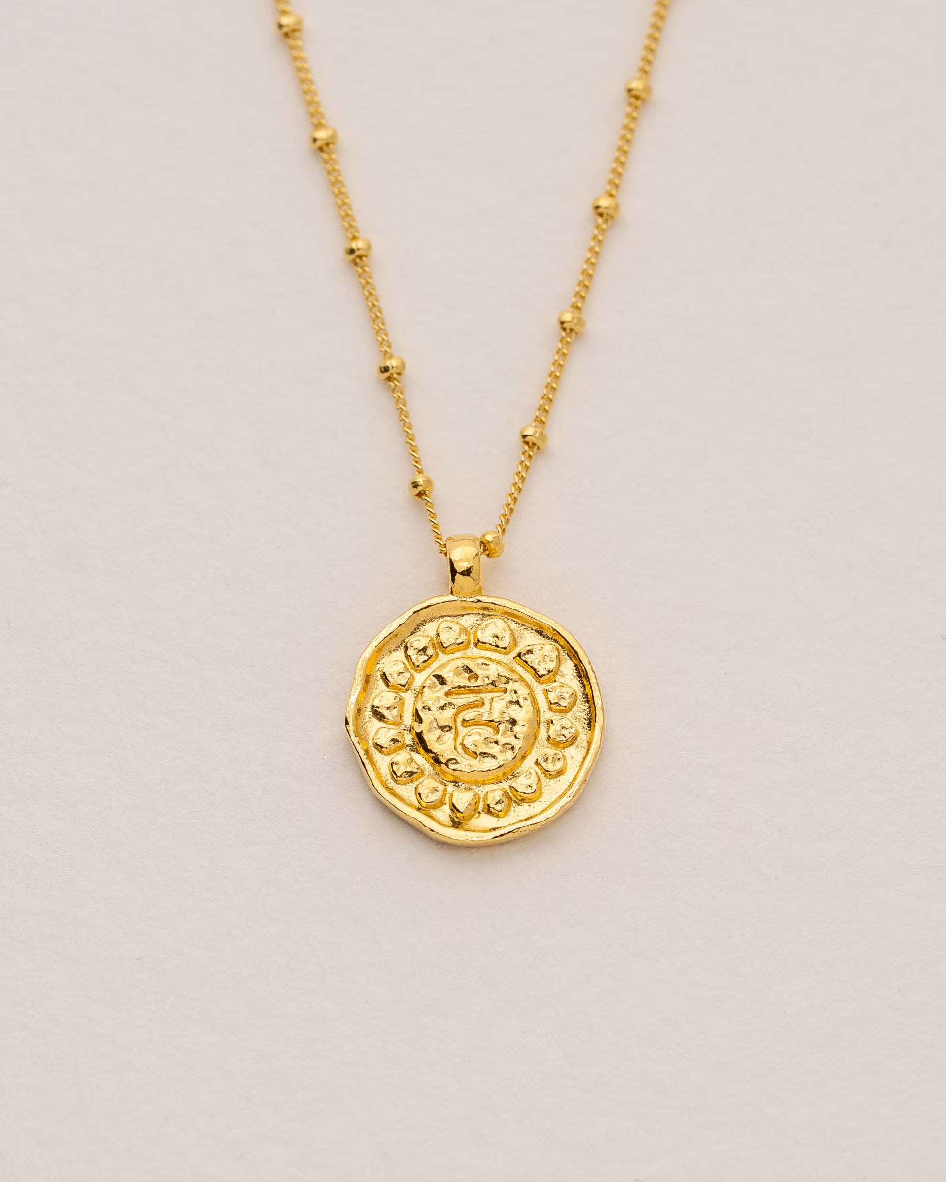 #Motiv_Vishuddha CHAKRA VISHUDDHA NECKLACE GOLD, goldene Halskette von ILUH, handgefertigt auf Bali – Spirituelles Produktfoto im minimalistischen Boho-Look von einer feinen Satellitenkette aus 18k vergoldetem 925er Silber mit einem runden Münzanhänger, der das originale Sanskrit-Zeichen für das Vishuddha-Halschakra, eingebettet in kunstvolle Lotusblütenblätter zur Stärkung des Energiezentrums, zeigt, flach präsentiert auf einem hellen, neutralen Büttenpapier-Untergrund.