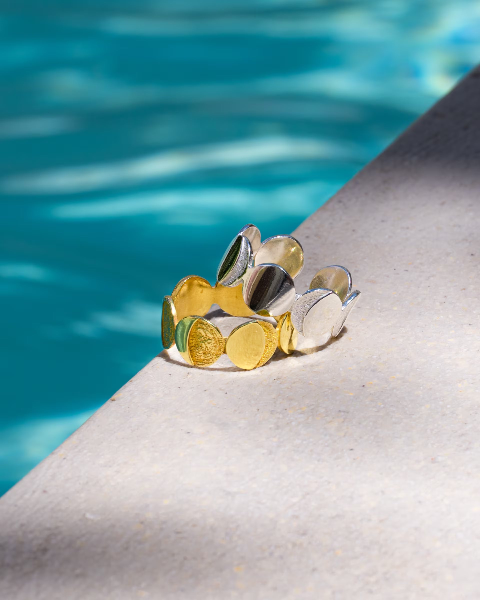 CHANDRA MOONPHASES RING, goldener und silberner Schmuck von ILUH, handgefertigt auf Bali – Stilvolles Produktfoto im sommerlichen Look, das zwei breite, offene Mondphasen-Ringe in der Bicolor-Variante aus 18k vergoldetem 925er Silber und purem Silber zeigt, wie diese dekorativ auf einem hellen Beckenrand vor türkisblau schimmerndem Poolwasser präsentiert werden.