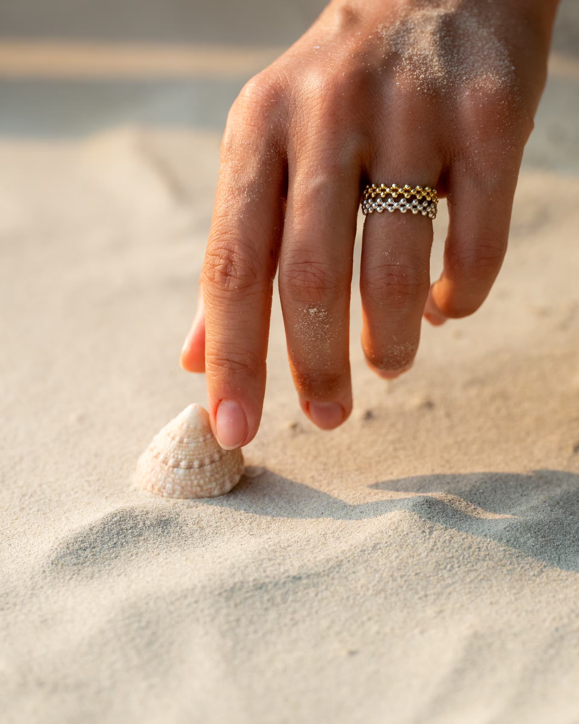 ERIS RING STACK, silberner und goldener Schmuck von ILUH, handgefertigt auf Bali – Detailreiche Nahaufnahme im maritimen Beach-Look von einer leicht mit Sand bedeckten Hand, die nach einer kleinen Muschel im hellen Strandsand greift und dabei eine stilvolle Kombination aus mehreren, übereinandergesteckten ERIS RINGEN in Bicolor-Optik aus 925er Silber und 18k Vergoldung zeigt.