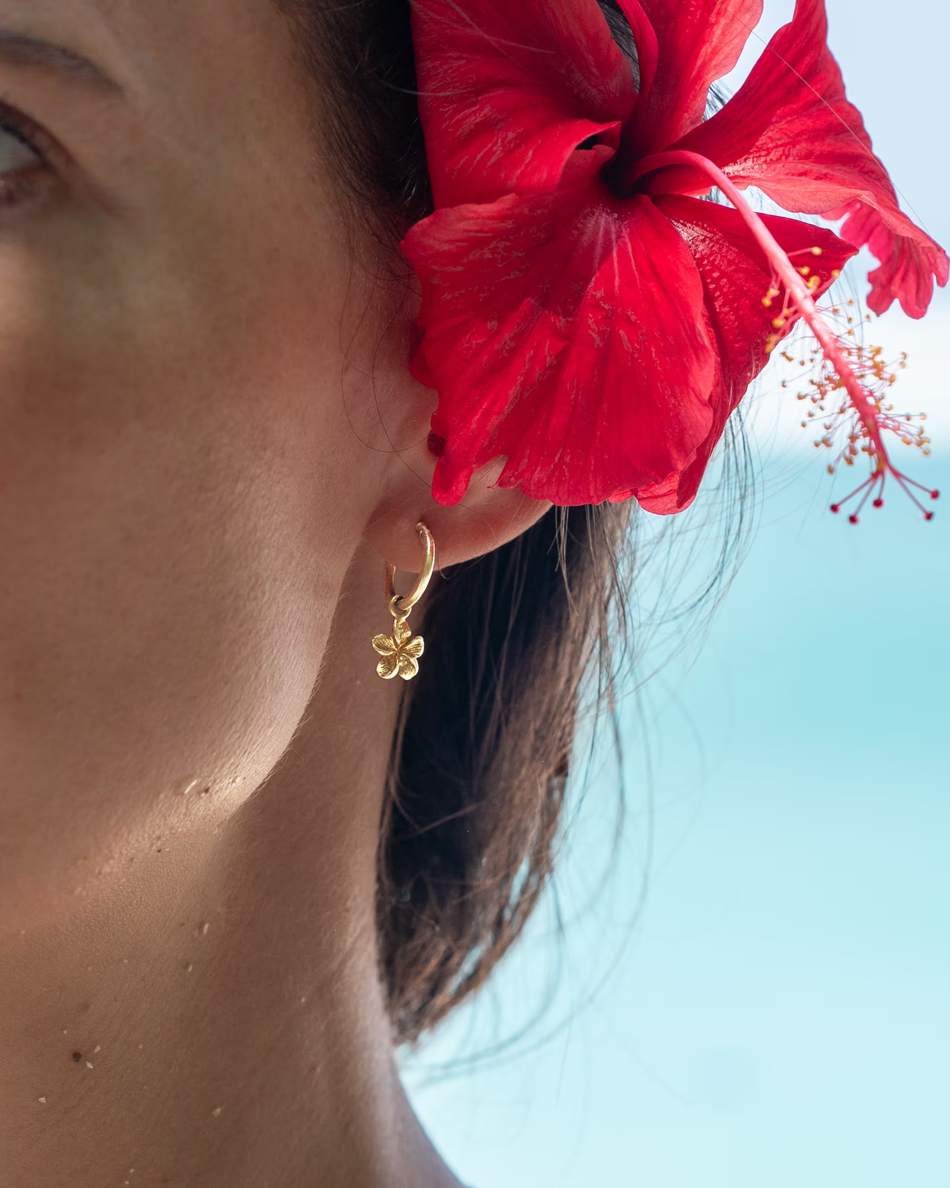 FRANGIPANI FLOWER OHRRINGE, kleine goldene Creolen von ILUH, handgefertigt auf Bali – Detailreiche Nahaufnahme im Profil von einer Frau am Strand mit einer leuchtend roten Hibiskusblüte im Haar, die den eleganten Ohrschmuck aus 18k vergoldetem 925er Silber mit zartem Blüten-Anhänger perfekt in Szene setzt.