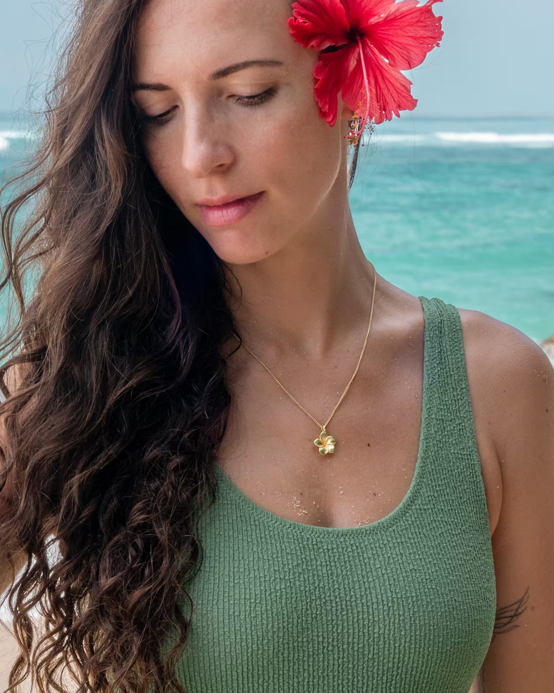 Stimmungsvolle Nahaufnahme im tropischen Beach-Look von einer Frau mit langen, lockigen braunen Haaren im salbeigrünen Rippstrick-Badeanzug am Strand, die eine leuchtend rote Hibiskusblüte im Haar trägt und den Blick senkt, während sie die auf Bali handgefertigte goldene FRANGIPANI KETTE mit einem filigranen Blüten-Anhänger aus 18k vergoldetem 925er Silber von der Schmuckmarke ILUH zeigt.