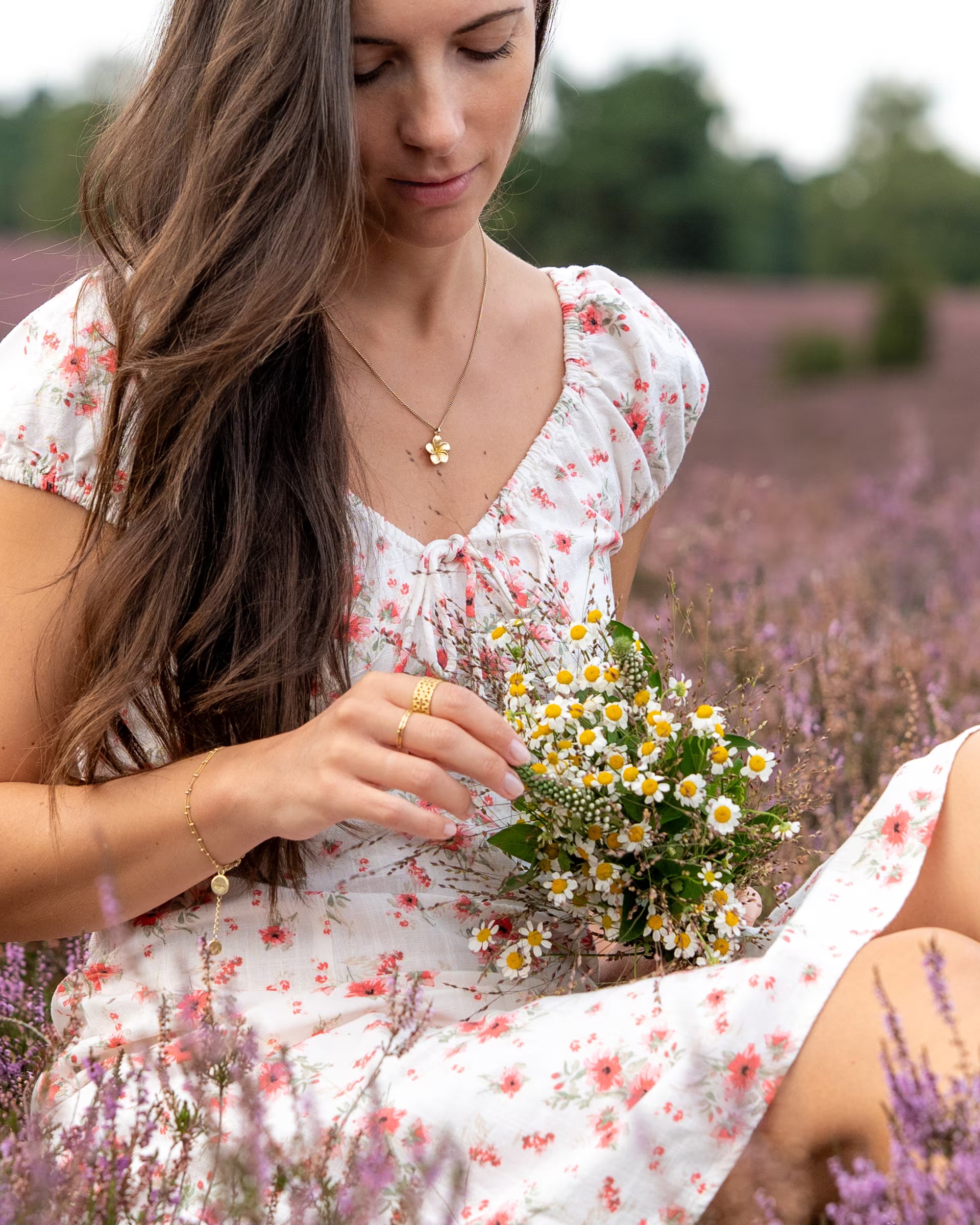 Romantische Nahaufnahme im sommerlichen Cottagecore-Stil von einer sitzenden Frau mit langen Haaren im floral gemusterten Kleid inmitten eines blühenden Heidekrautfeldes, die einen Strauß Wildblumen (Kamille) hält und eine Kombination aus auf Bali handgefertigtem goldenem Schmuck – bestehend aus der FRANGIPANI KETTE mit Blüten-Anhänger, einem feinen Gliederarmband sowie Ringen aus 18k vergoldetem 925er Silber von der Schmuckmarke ILUH – trägt.
