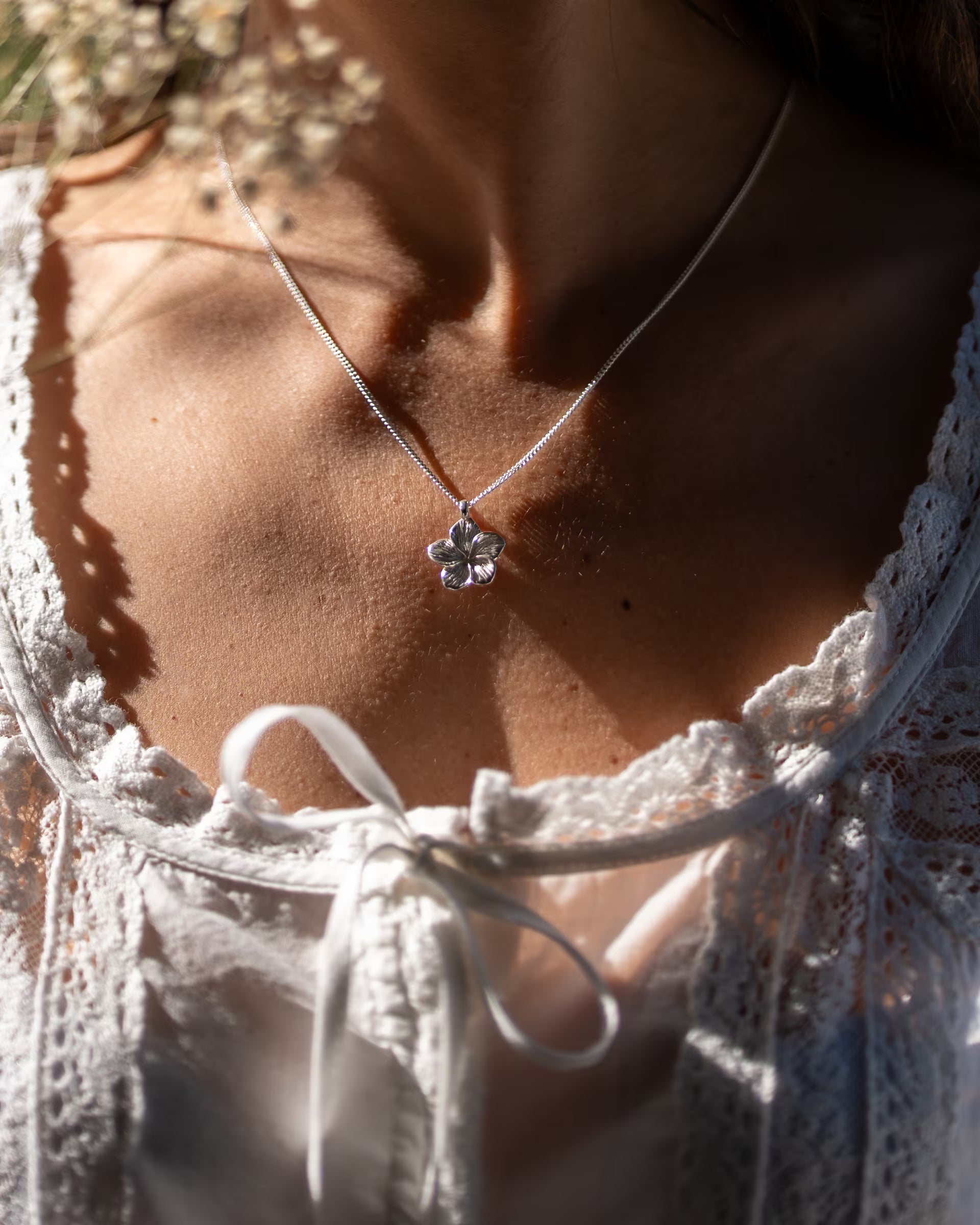 FRANGIPANI FLOWER KETTE, silberne Halskette von ILUH, handgefertigt auf Bali – Romantische Nahaufnahme im verspielten Cottagecore-Look von dem Dekolletee einer Frau in einer weißen, feinen Spitzenbluse mit Schleifendetail, auf deren Haut der filigrane Frangipani-Blütenanhänger aus 925er Silber in einem weichen Spiel aus Licht und Schatten erstrahlt.