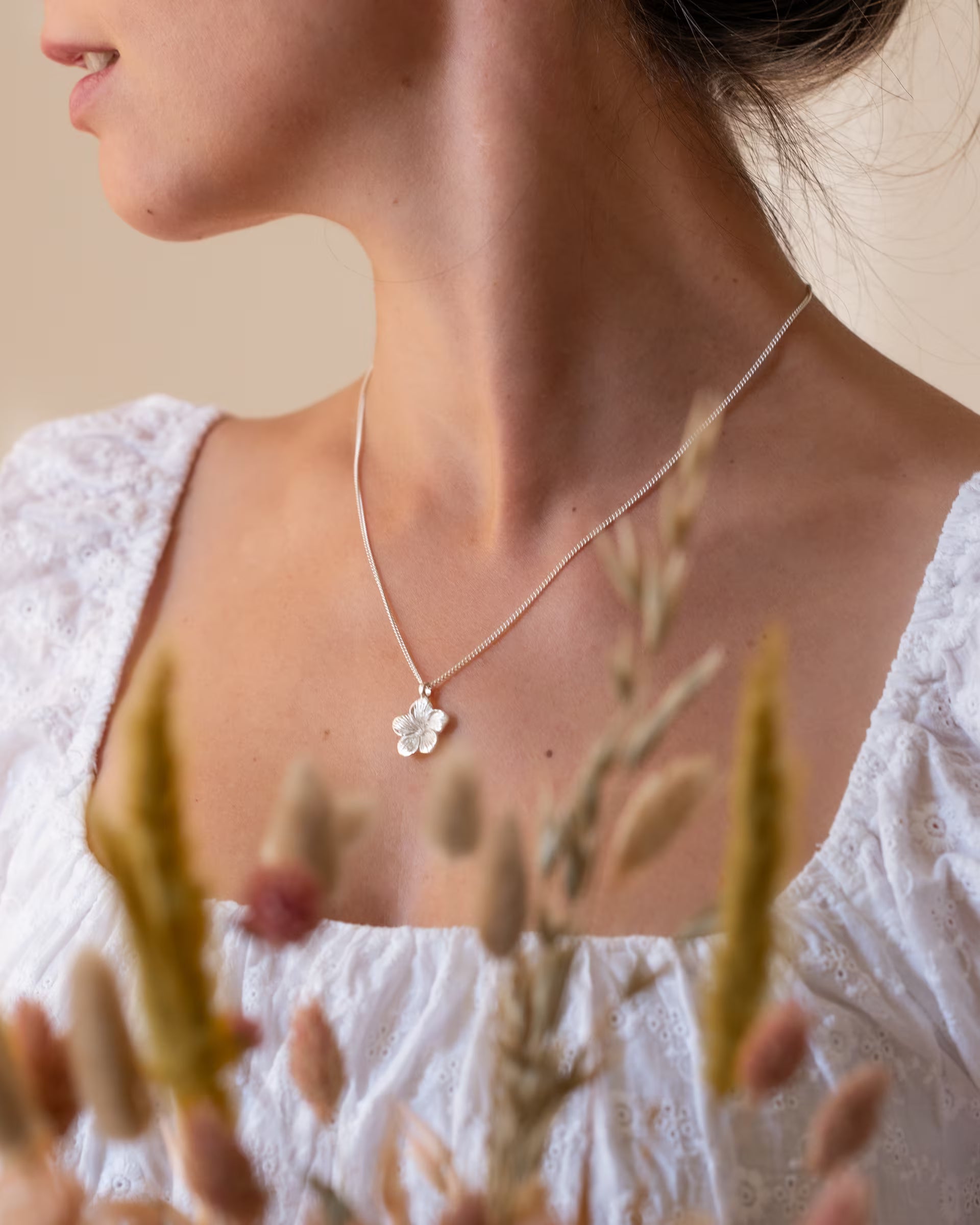FRANGIPANI FLOWER KETTE, silberne Halskette von der Marke ILUH, handgefertigt auf Bali – Elegante, verträumte Nahaufnahme von dem Dekolleté einer wegschauenden Frau in einer weißen Spitzenbluse, die eine feine Halskette aus 925er Silber mit einem detailreich gestalteten Frangipani-Blütenanhänger trägt, während im Vordergrund zarte Trockenblumen sanft ins Bild ragen.