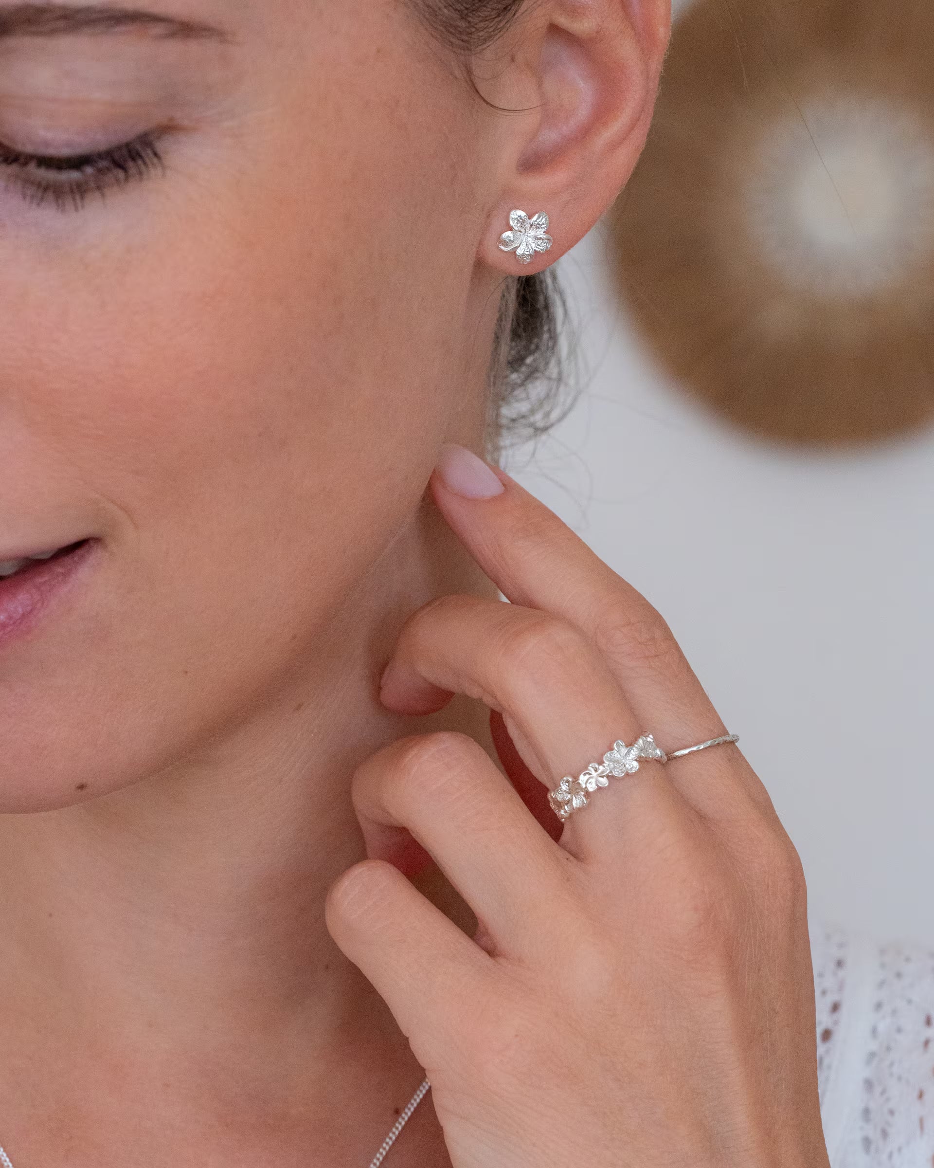 Auf Bali handgefertigter silberner Frangipani Flower Ring und passende Frangipani Ohrstecker mit Blüten-Motiv aus 925er Silber von der Schmuckmarke ILUH, getragen im modernen Boho-Stil von einer Frau vor einer Wand mit Makramee- und Bast-Deko.