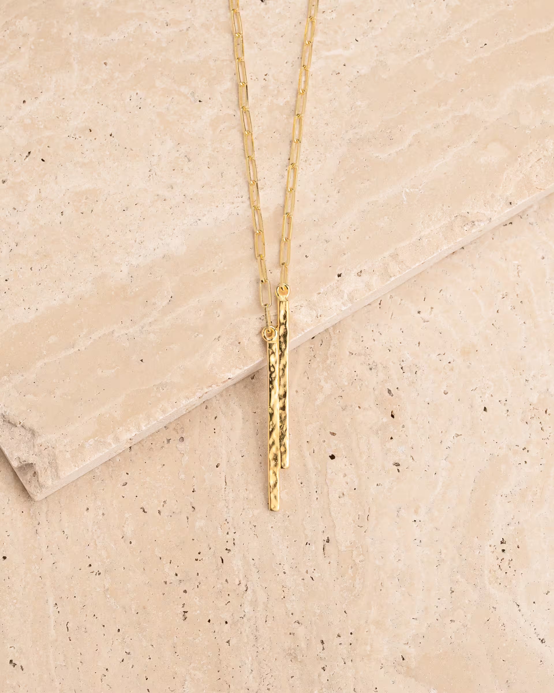 FUSE HAMMERED NECKLACE GOLD, goldene Halskette von ILUH, handgefertigt auf Bali – Minimalistisches Produktfoto im modernen Boho-Look von einer feinen Gliederkette aus 18k vergoldetem 925er Silber mit zwei unterschiedlich langen, schmalen Stabanhängern, die eine markante, gehämmerte Struktur aufweisen, elegant präsentiert auf einem hellen, gemaserten Naturstein-Untergrund.