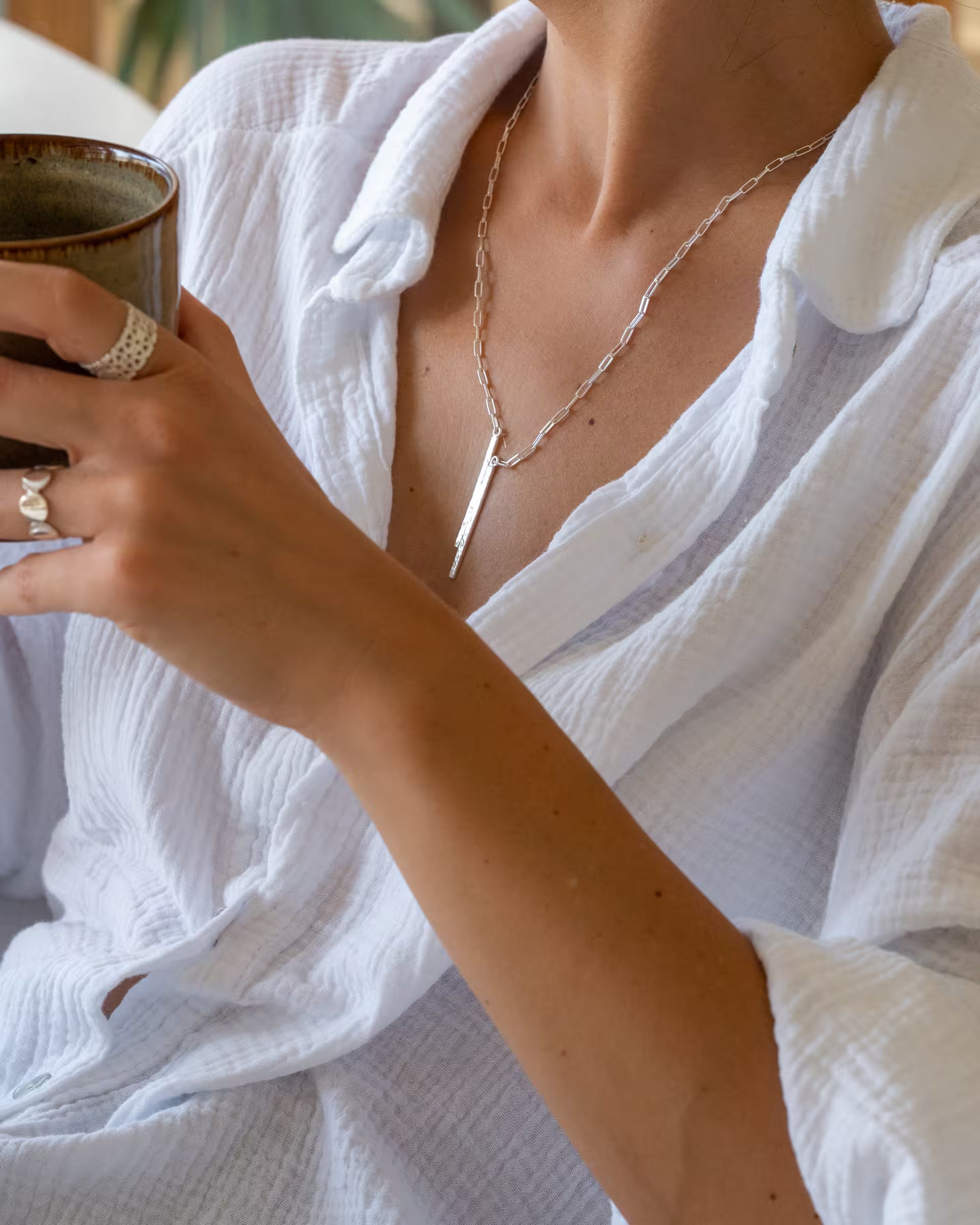 Nahaufnahme im entspannten Alltags-Look von einer jungen Frau in einer weißen Musselin-Bluse, die eine Kaffeetasse hält und eine Kombination aus auf Bali handgefertigtem silbernem Schmuck – darunter die FUSE PAPERCHAIN KETTE mit einem länglichen, gehämmerten Stab-Anhänger an einer Gliederkette, den breiten, offenen ENDLESS LOVE RING mit Herz-Muster sowie den offenen CHANDRA MOONPHASES Ring – aus 925er Silber von der Schmuckmarke ILUH trägt.