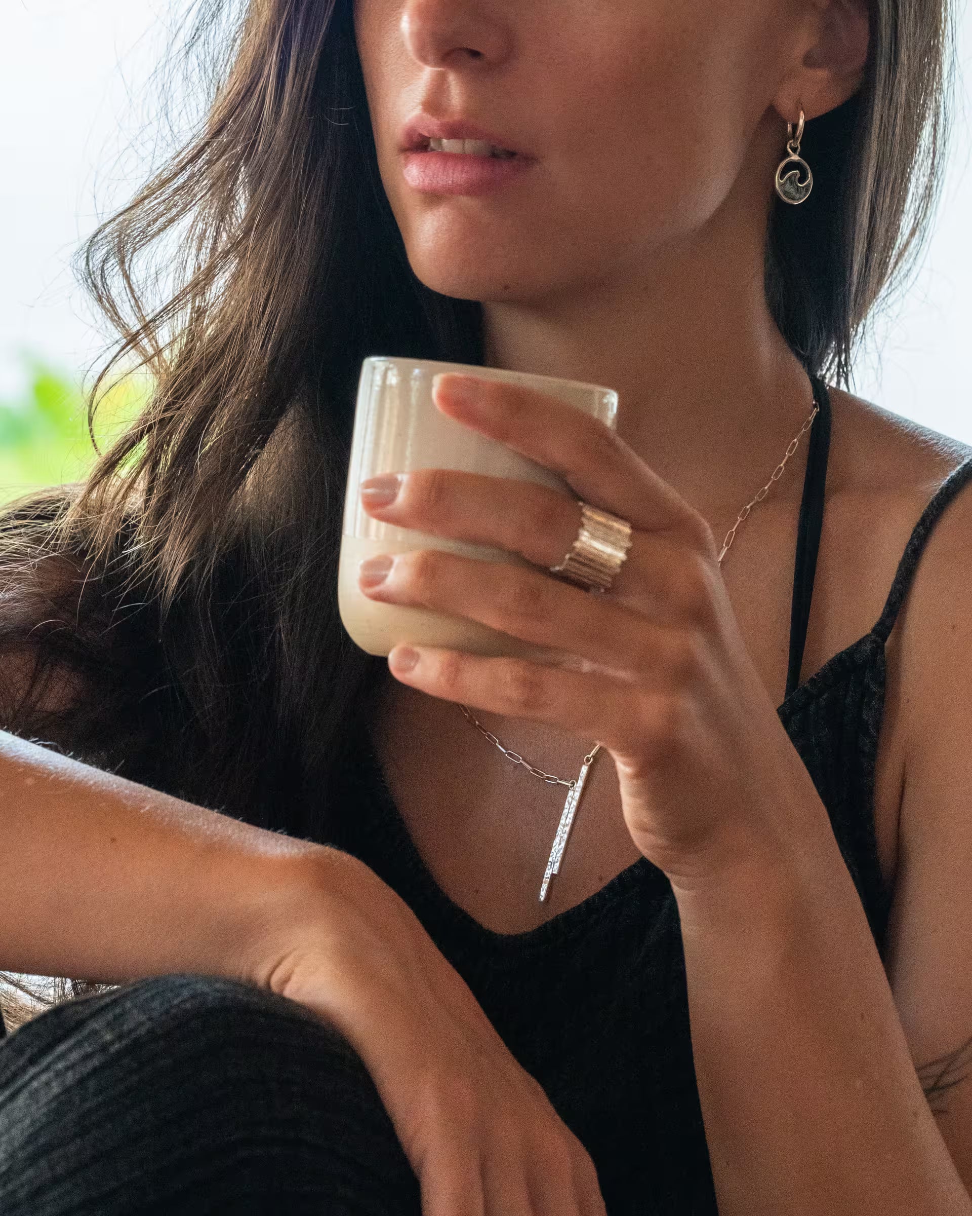 Junge Frau im lässigen Sommer-Look in einem Café, sie trägt handgefertigte LAJU Creolen-Ohrringe aus 925er Silber mit Abalone-Muschel, kombiniert mit der FUSE Paperchain-Kette und einem breiten, gehämmerten FUSE Ring vom Schmucklabel ILUH, während sie eine weiße Kaffeetasse in der Hand hält.