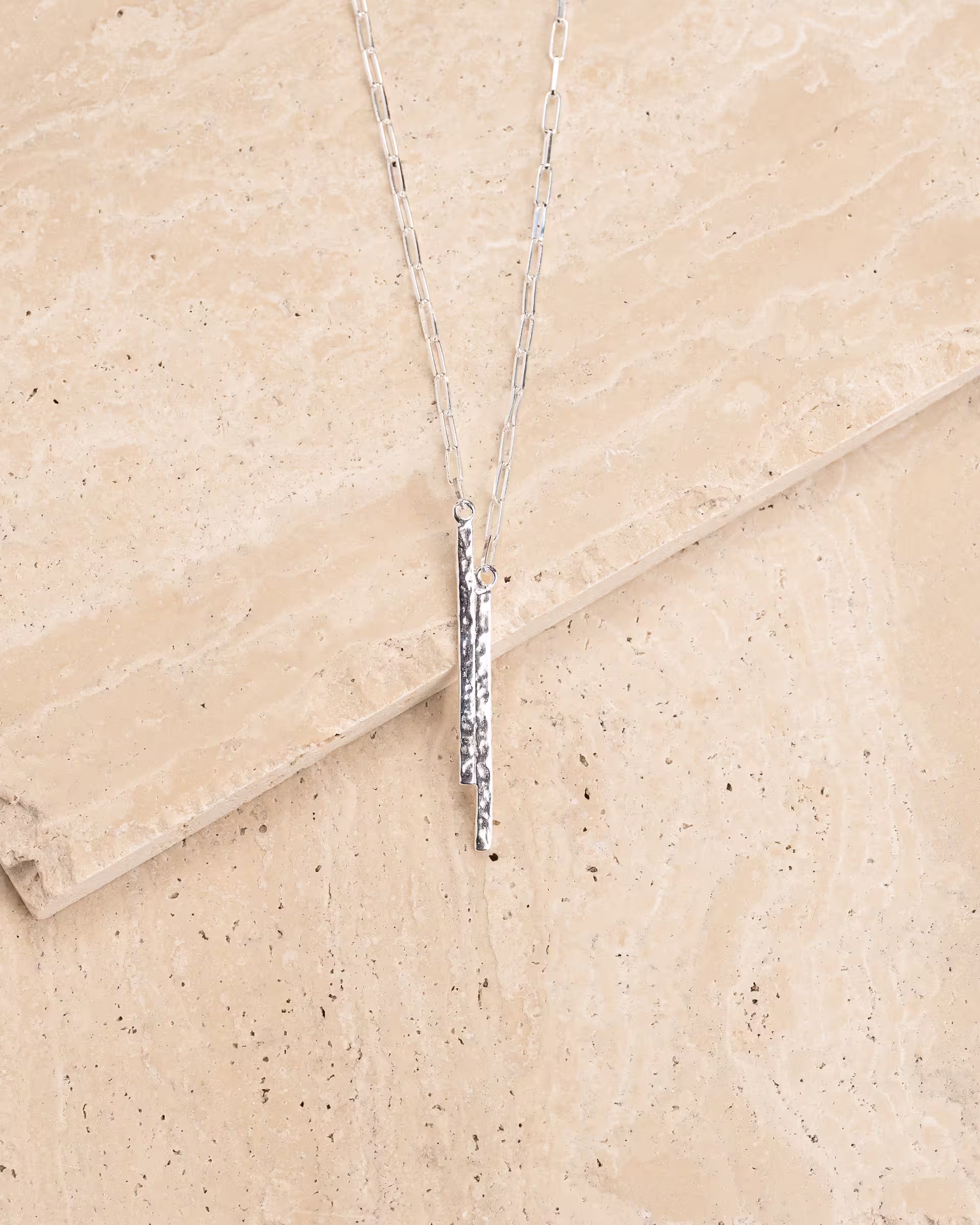 FUSE HAMMERED NECKLACE SILBER, silberne Halskette von ILUH, handgefertigt auf Bali – Minimalistisches Produktfoto im modernen Boho-Look von einer feinen Paperchain Gliederkette aus 925er Silber mit zwei unterschiedlich langen, schmalen Stabanhängern, die eine markante, gehämmerte Struktur aufweisen, elegant präsentiert auf einem hellen, gemaserten Naturstein-Untergrund.