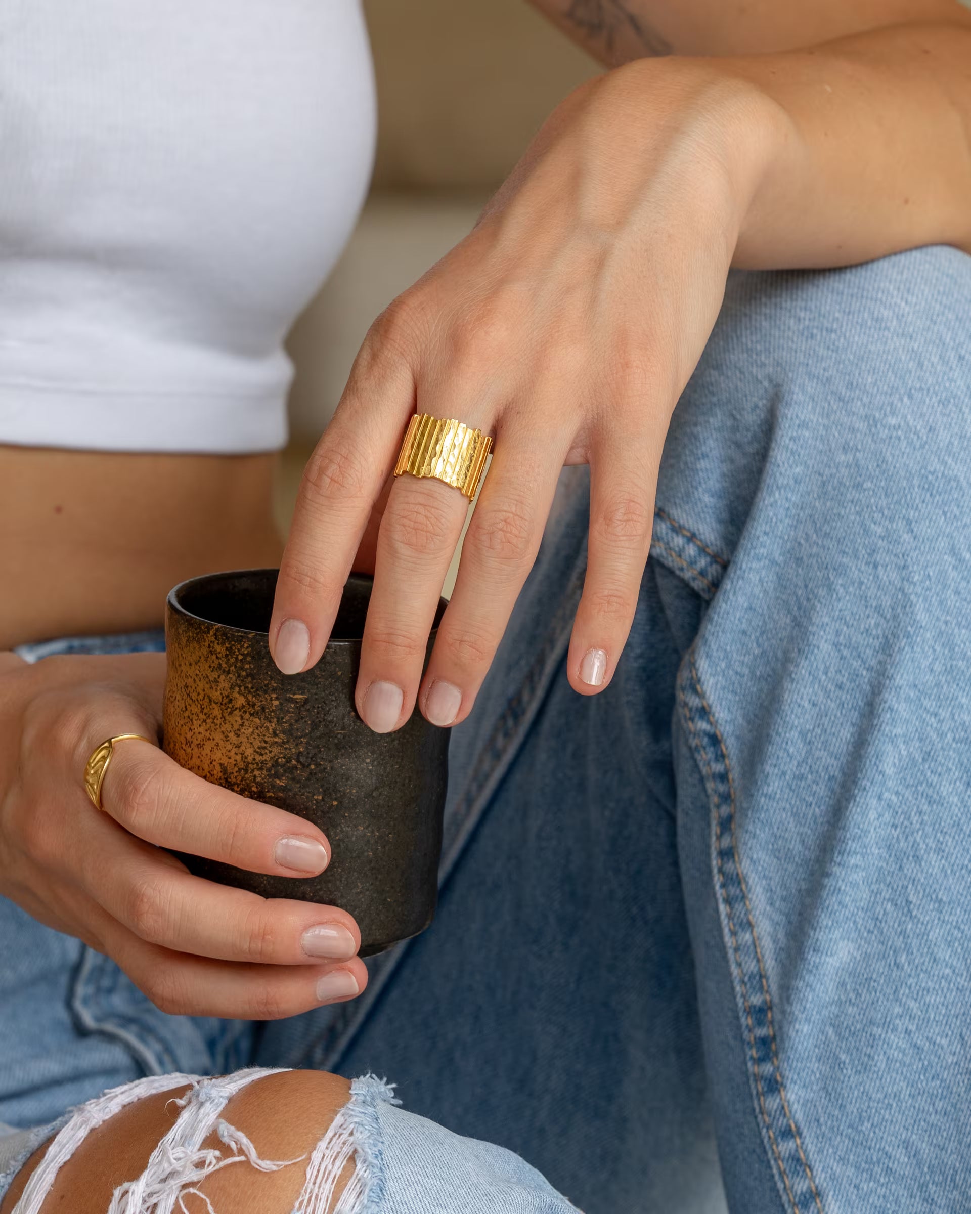 Breiter, gehämmerter FUSE Statement-Ring in Gold von der Schmuckmarke ILUH, getragen an der Hand einer Frau mit einer Keramiktasse und Destroyed-Jeans.