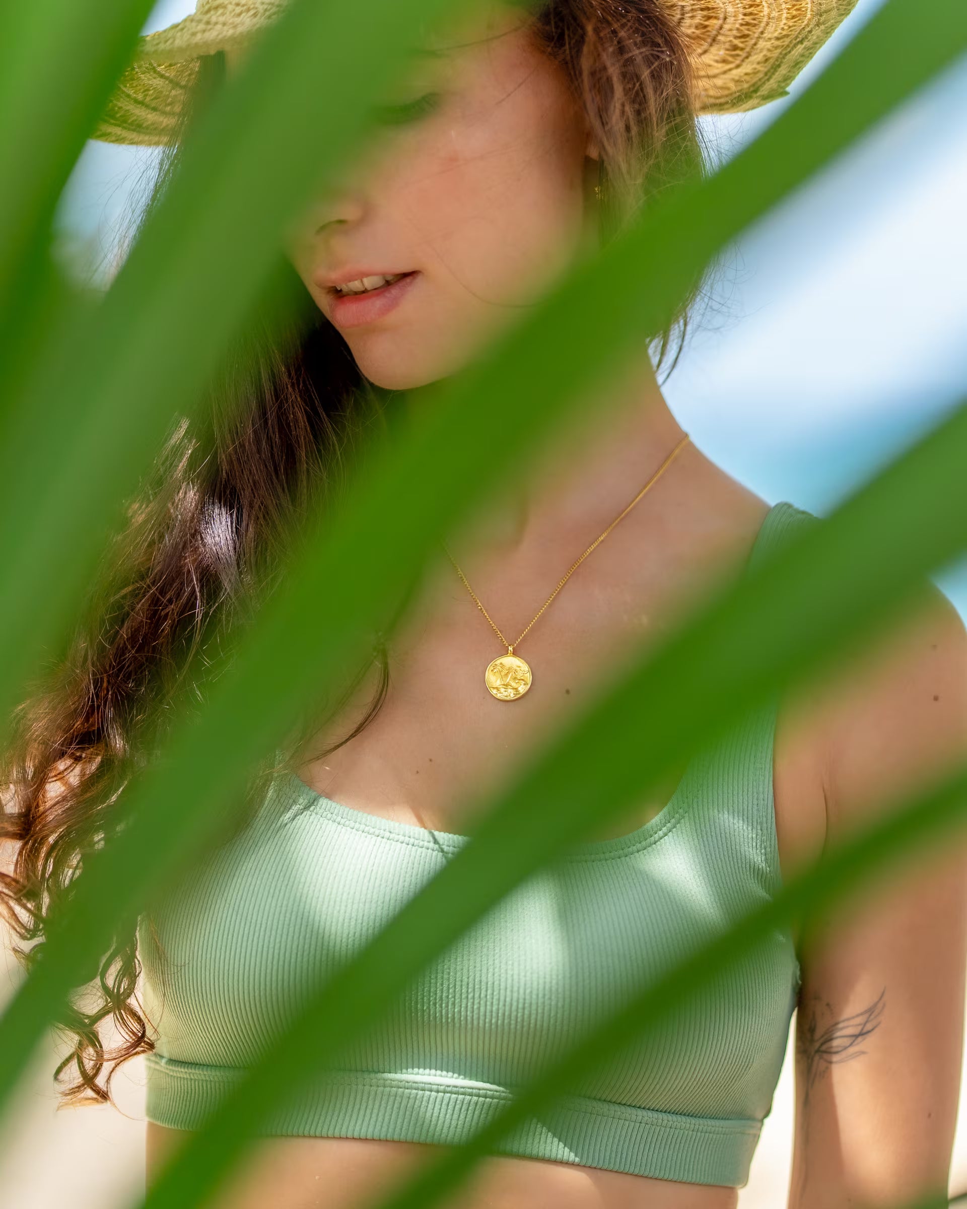 Nahaufnahme im sommerlichen Beach-Look von einer Frau im salbeigrünen Bikini-Top und Strohhut, die die auf Bali handgefertigte goldene GILI PARADISE ISLAND Kette mit rundem Münz-Anhänger aus 18k vergoldetem 925er Silber von der Schmuckmarke ILUH trägt, fotografiert durch grüne Palmenblätter.