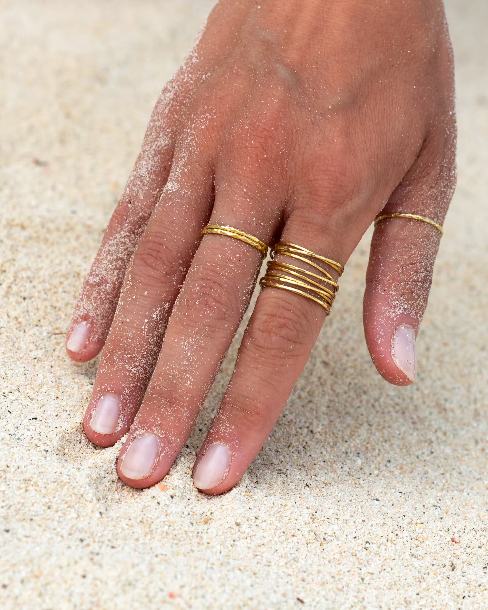 Nahaufnahme einer sandigen Hand im Strandsand, die den auf Bali handgefertigten goldenen AURA Ring im Stacking-Look sowie den feinen SARI Ring aus 18k vergoldetem 925er Silber von der Schmuckmarke ILUH als Ring-Kombination im sommerlichen Beach-Look trägt.
