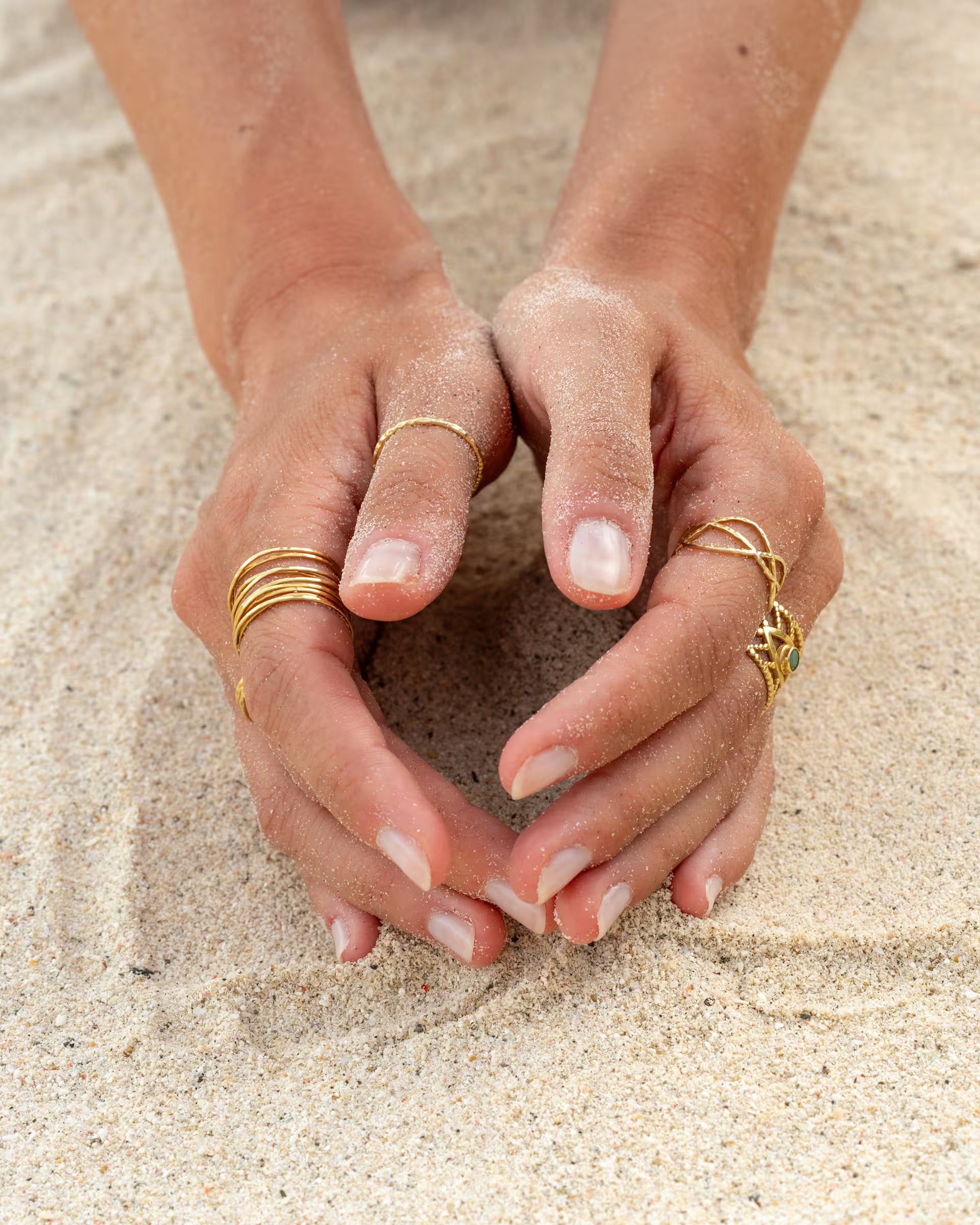 Handgefertigtes Set aus goldenen AURA, SARI, LOTUS und INTUITION Ringen der Schmuckmarke ILUH, kunstvoll präsentiert an den Händen einer jungen Frau, die ihre Finger sanft in den hellen Sand am Strand eingräbt; ein detailreicher Beach-Look auf Bali mit Fokus auf filigrane Goldringe.