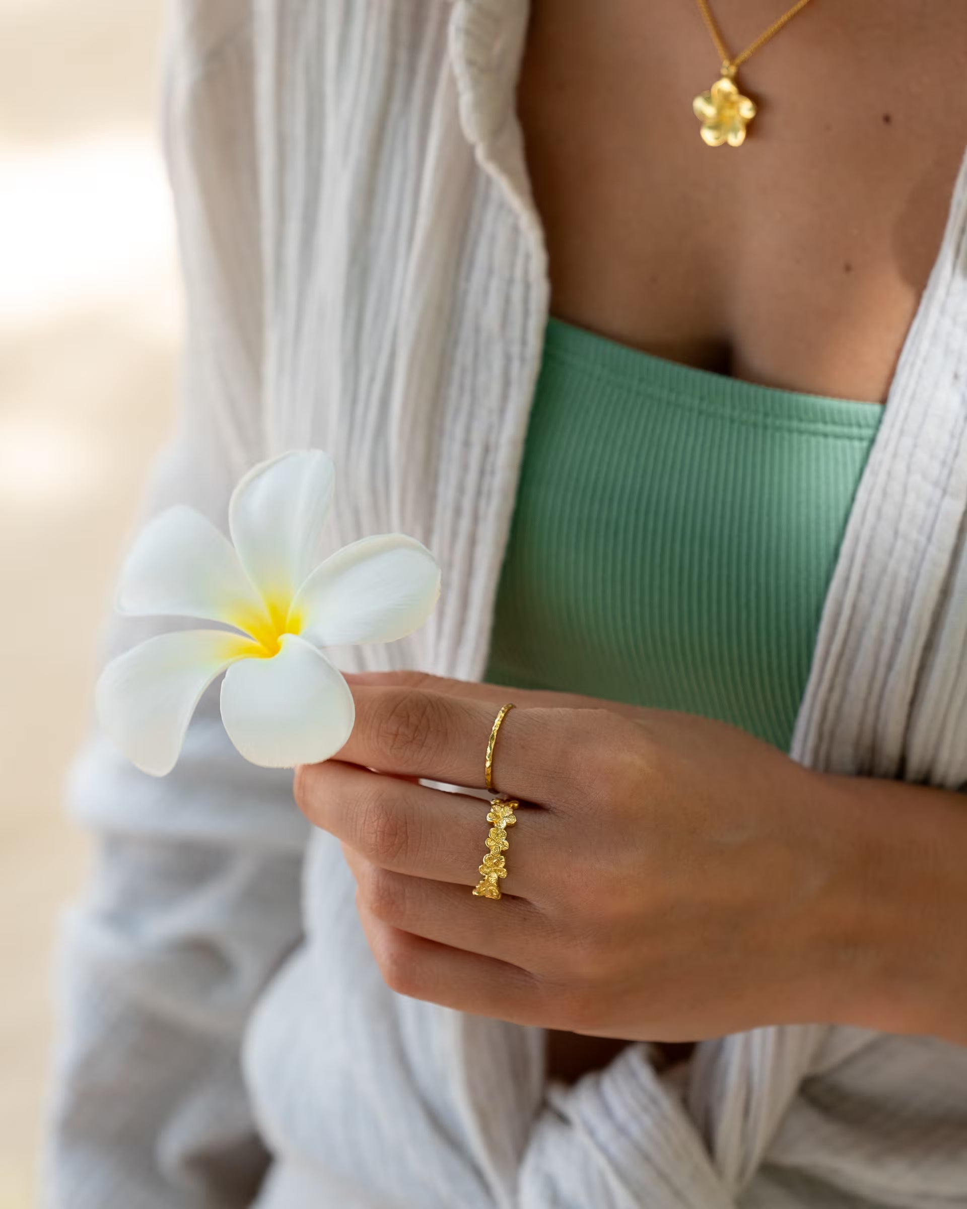 Nahaufnahme im sommerlichen Boho-Look von einer Frau in einer hellen Musselin-Bluse und salbeigrünen Top, die eine weiße Frangipani-Blüte hält und eine Kombination aus auf Bali handgefertigtem goldenem Schmuck – bestehend aus der FRANGIPANI KETTE mit Blüten-Anhänger, dem filigranen FRANGIPANI FLOWER RING aus aneinandergereihten Blüten sowie dem feinen, gehämmerten SARI STACKING RING – aus 18k vergoldetem 925er Silber von der Schmuckmarke ILUH trägt.