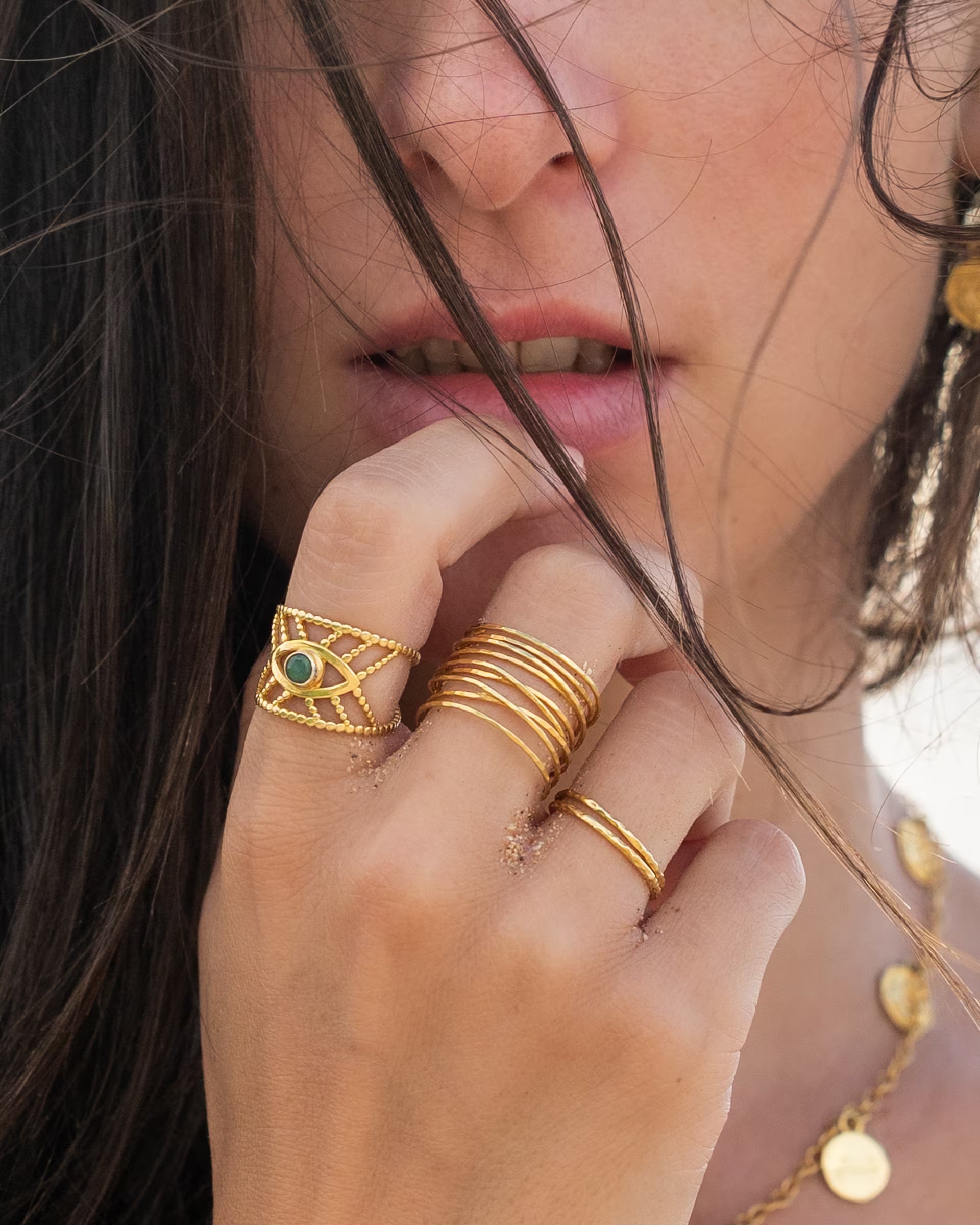Junge Frau im Beach-Look trägt mehrere goldene Ringe, darunter einen breiten Statement-Ring mit grünem Smaragd Edelstein im Dritten-Auge-Design zur Stärkung der Intuition, kombiniert mit dem vergoldeten AURA- und SARI-Ring von der Schmuckmarke ILUH.