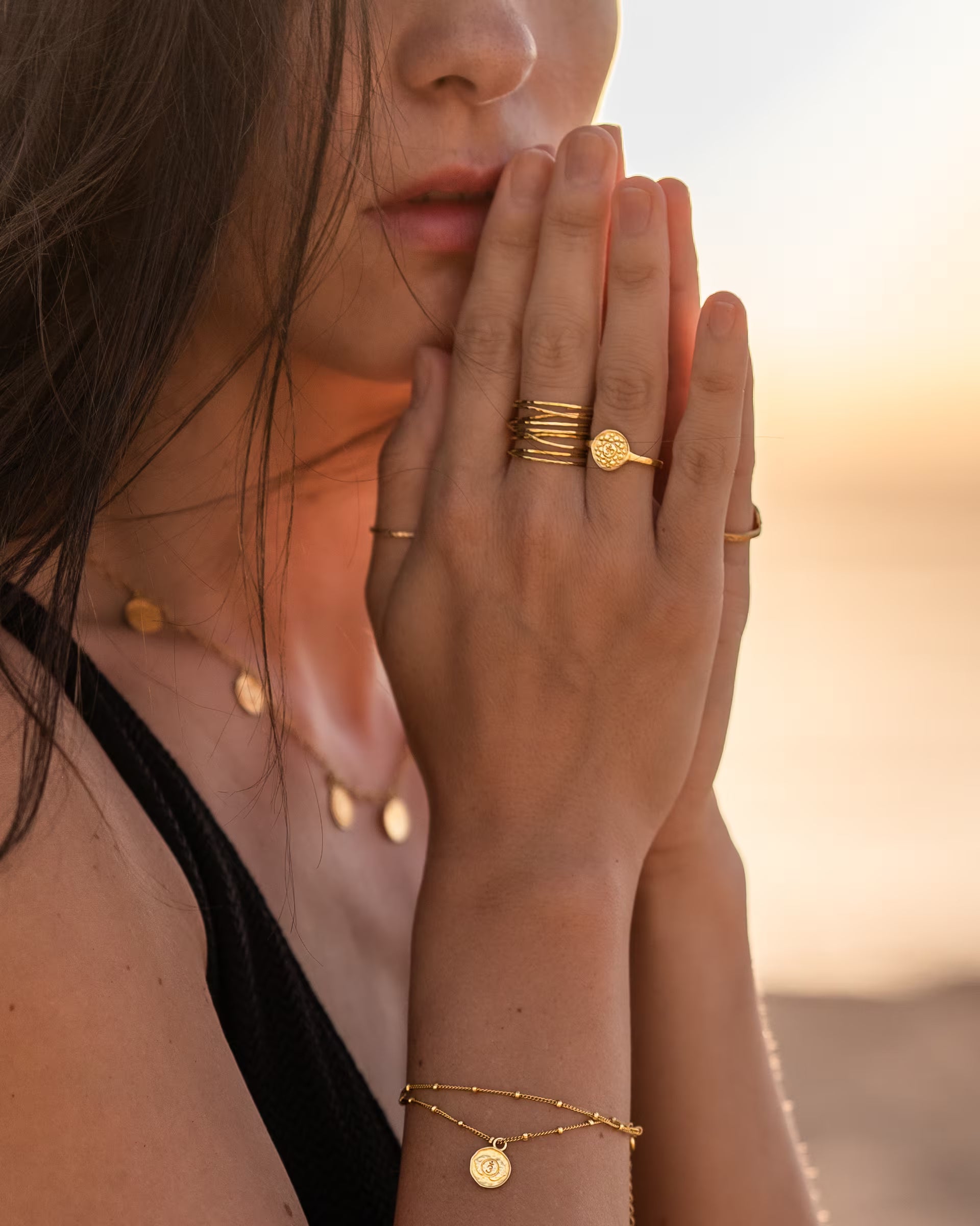Nahaufnahme im stimmungsvollen Beach-Look von einer Frau im schwarzen Top, die ihre Hände sanft an das Gesicht legt und eine Kombination aus auf Bali handgefertigtem goldenem Schmuck – darunter die PRANA CHAKRA KETTE mit allen sieben Chakra-Motiven, das passende CHAKRA ARMBAND, den AURA RING im Stacking-Look sowie CHAKRA RINGE – aus 18k vergoldetem 925er Silber von der Schmuckmarke ILUH im warmen Licht des Sonnenuntergangs trägt.
