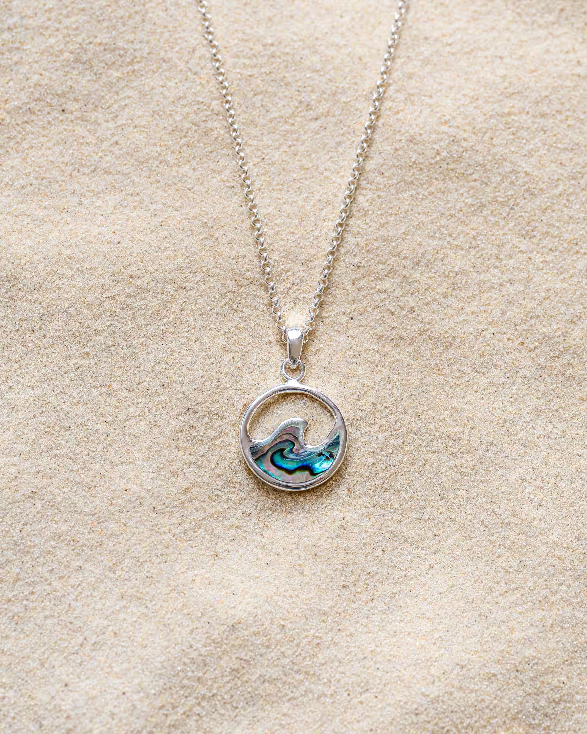 LAJU ABALONE NECKLACE SILBER, silberne Halskette von ILUH, handgefertigt auf Bali – Elegantes Produktfoto im maritimen Beach-Look von einer feinen Halskette aus 925er Silber mit einem runden, offenen Anhänger, der eine geschwungene Meereswelle und eine intensiv blau-grün schimmernde Einlage aus echter Abalone-Muschel zeigt, flach präsentiert im feinen Sandstrand.
