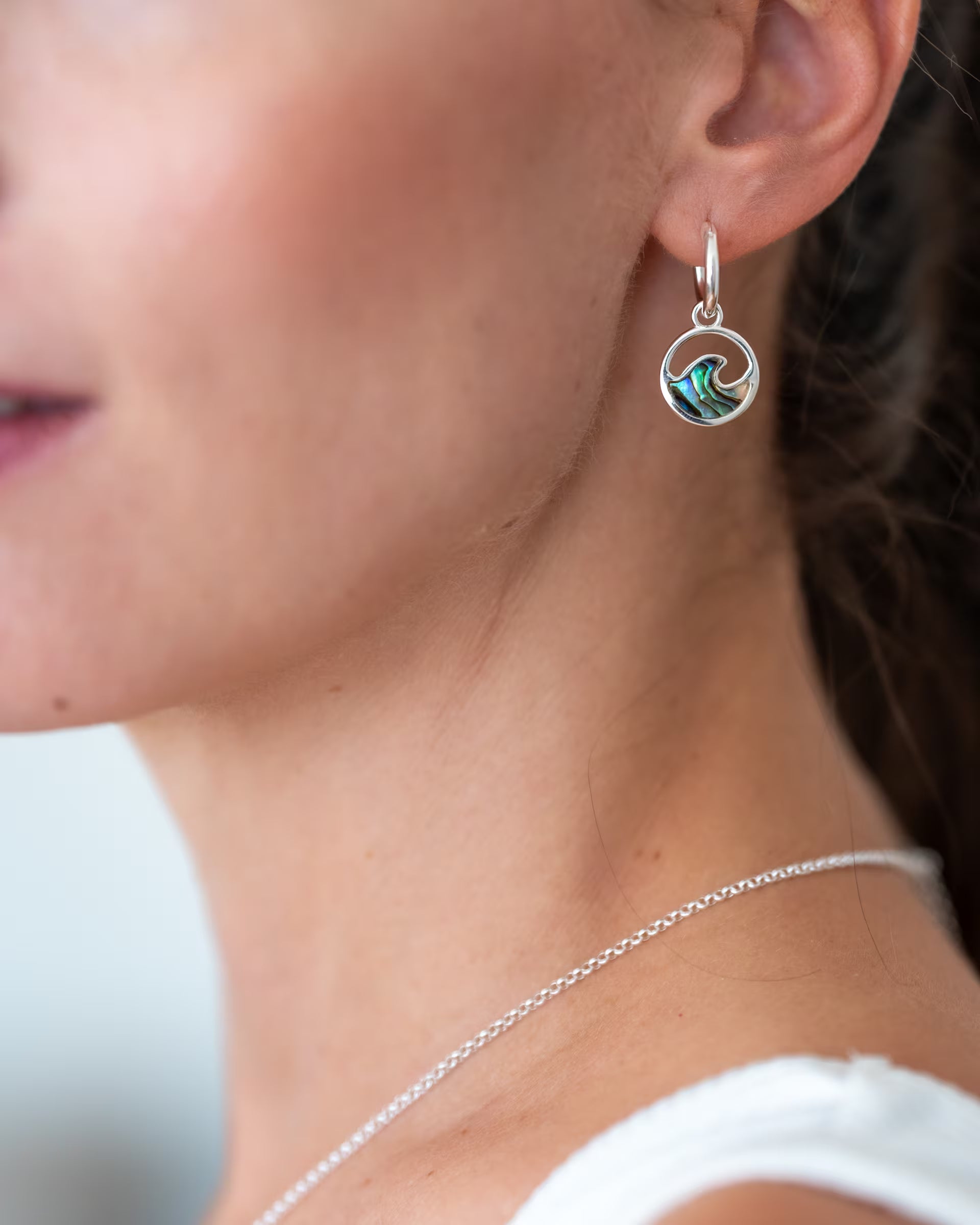 Nahaufnahme im minimalistisch-sommerlichen Look von einer Frau im Profil, die die auf Bali handgefertigten silbernen LAJU WAVE ABALONE CREOLEN mit einem runden Wellen-Anhänger aus schimmernder Abalone-Muschel sowie eine feine Gliederkette aus 925er Silber von der Schmuckmarke ILUH trägt.