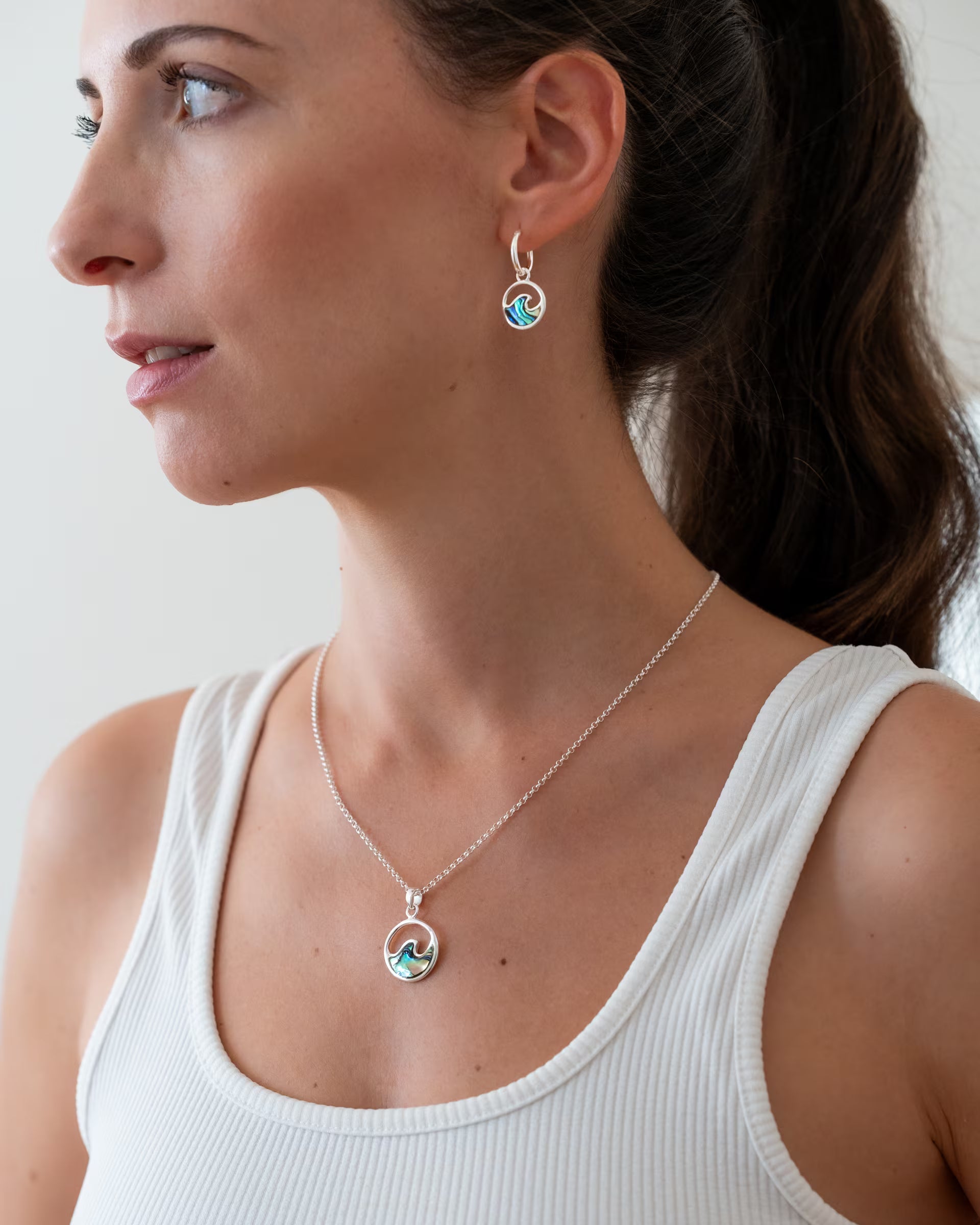 Nahaufnahme im minimalistisch-sommerlichen Look von einer Frau im weißen Rippstrick-Oberteil im Profil, die eine Kombination aus auf Bali handgefertigtem silbernem Schmuck – bestehend aus der LAJU WAVE ABALONE KETTE und den passenden LAJU WAVE ABALONE CREOLEN, jeweils mit runden Wellen-Anhängern aus blaugrün schimmernder Abalone-Muschel – aus 925er Silber von der Schmuckmarke ILUH trägt.