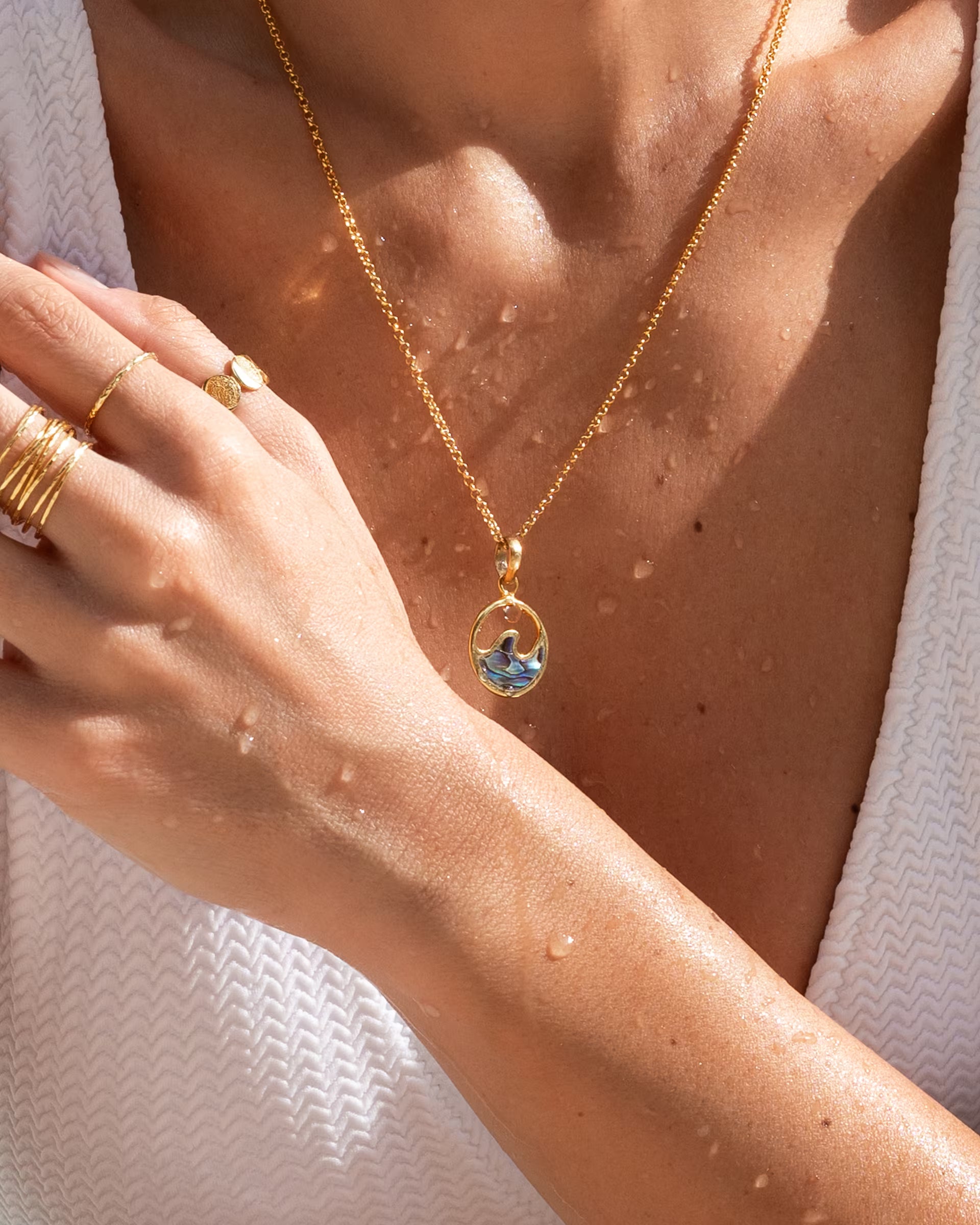 Auf Bali handgefertigte goldene LAJU Wave Abalone Kette mit einem runden Wellen-Anhänger aus Paua-Muschel und mehrere goldene Ringe aus 18k vergoldetem 925er Silber von der Schmuckmarke ILUH, getragen von einer Frau im weißen Oberteil mit Wassertropfen auf der Haut.