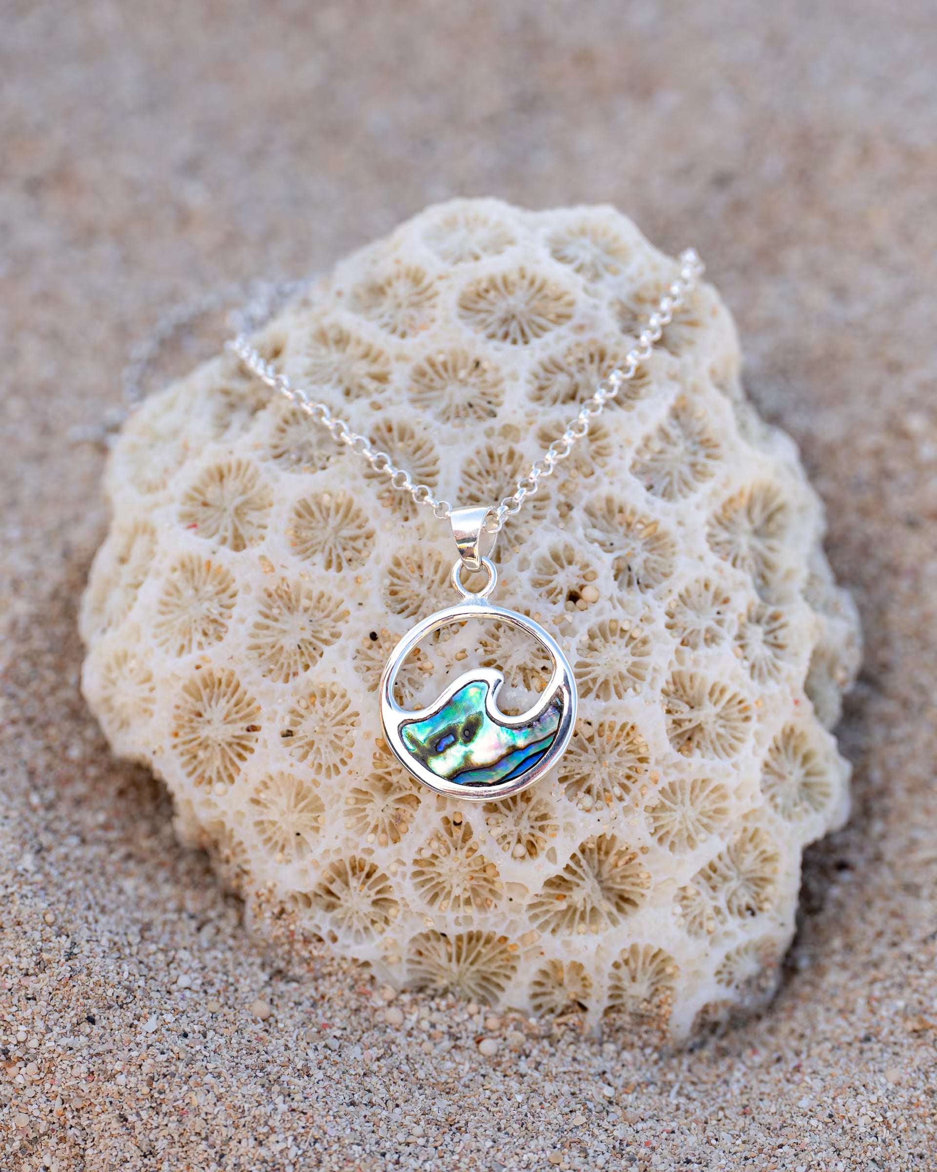 Handgefertigte LAJU Wellen-Halskette aus 925er Silber mit rundem Anhänger aus schimmernder Abalone-Muschel von der Schmuckmarke ILUH, elegant dekoriert auf einer hellen Koralle im Sand am Strand.