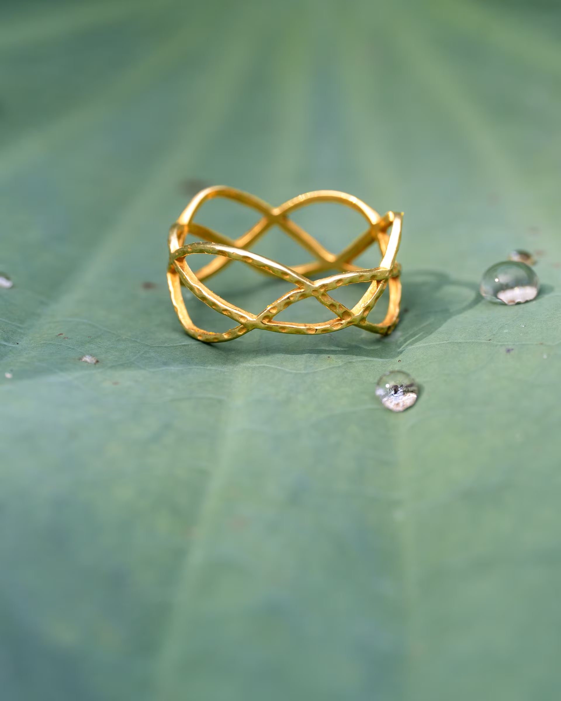 Produktfoto im naturverbundenen Stil von dem auf Bali handgefertigten goldenen LOTUS RING mit geschwungenem, geflochtenem Design aus 18k vergoldetem 925er Silber von der Schmuckmarke ILUH, platziert auf einem großen, grünen Lotusblatt mit feinen Wassertropfen.