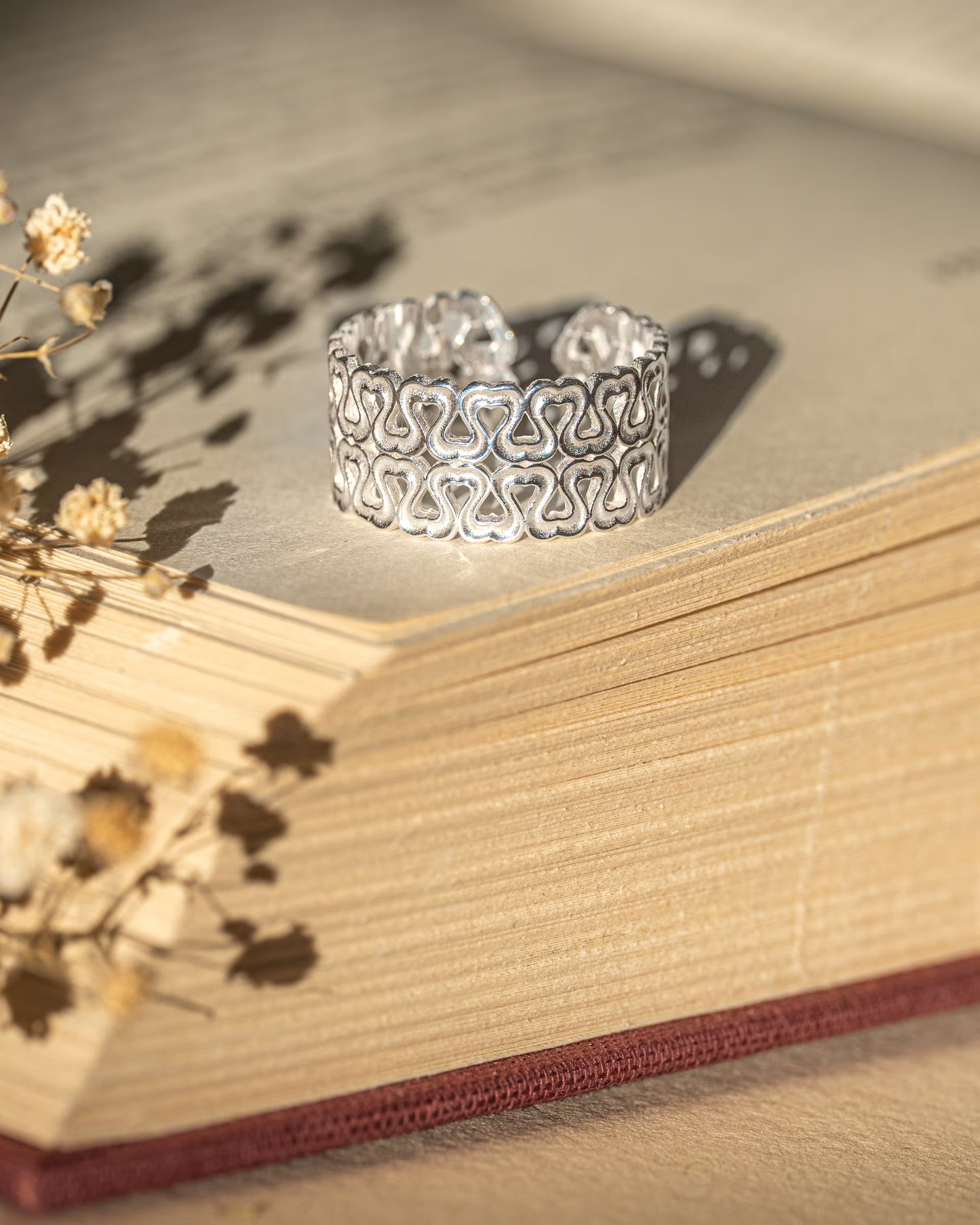 LOVE HEARTS RING SILBER, breiter silberner Ring von ILUH, handgefertigt auf Bali – Elegantes Produktfoto im romantischen Vintage-Look von einem breiten, offenen Ring aus 925er Silber mit einem filigranen, durchbrochenen Muster aus aneinandergereihten Herzen, der dekorativ auf den Buchseiten eines alten, aufgeschlagenen Buches neben zarten Trockenblumen präsentiert wird.